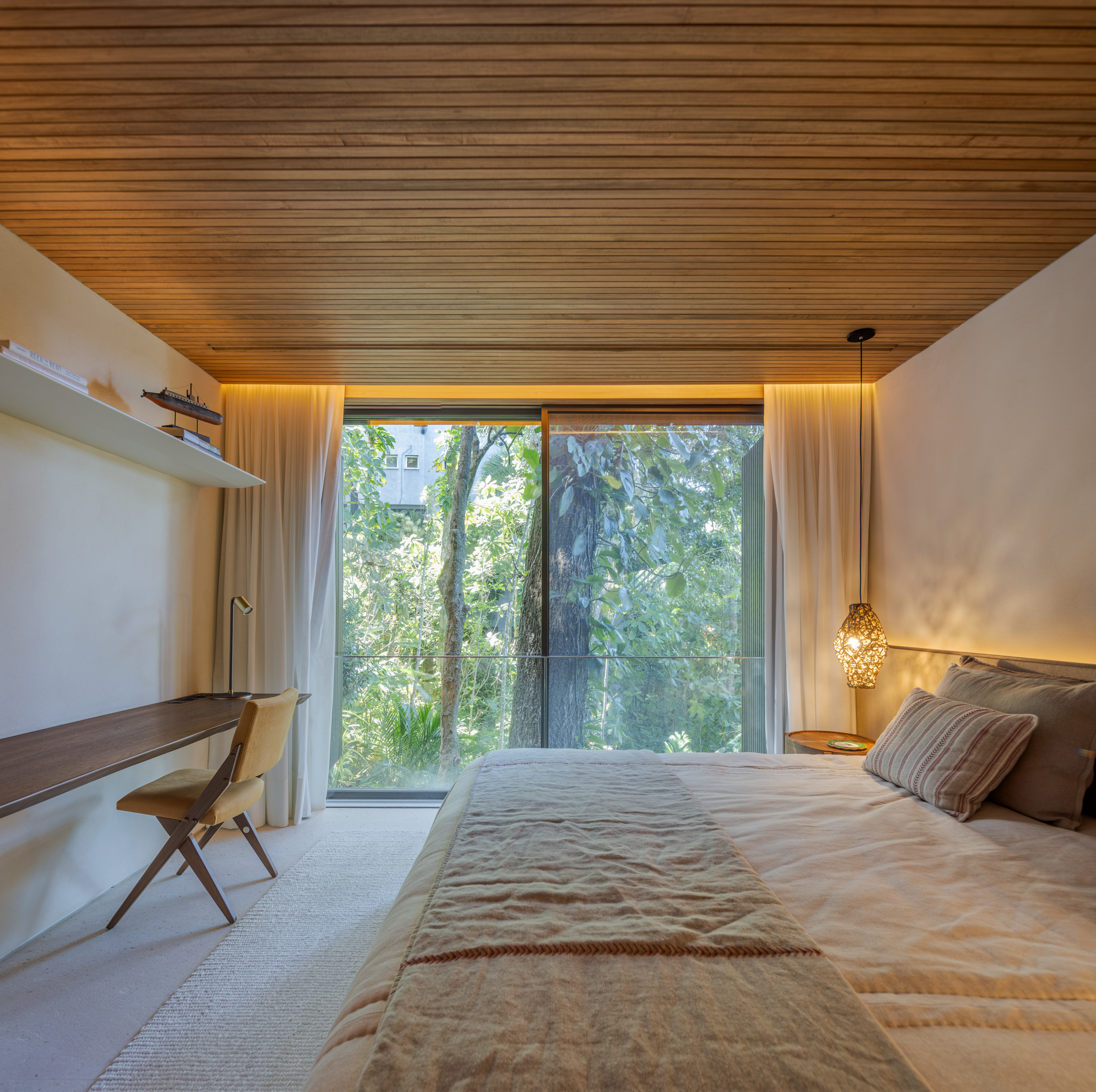 Bedroom with wooden ceiling, large window overlooking trees, bed with beige bedding, desk with chair, shelf, and pendant light on the right side—all thoughtfully arranged in a comfortable Passive House setting.