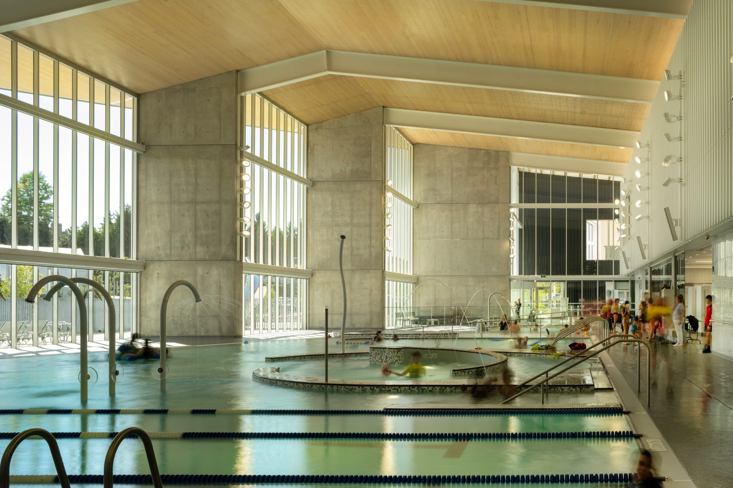 Indoor swimming pool with lap lanes, leisure pool, and large windows letting in natural light; people are swimming and walking around.