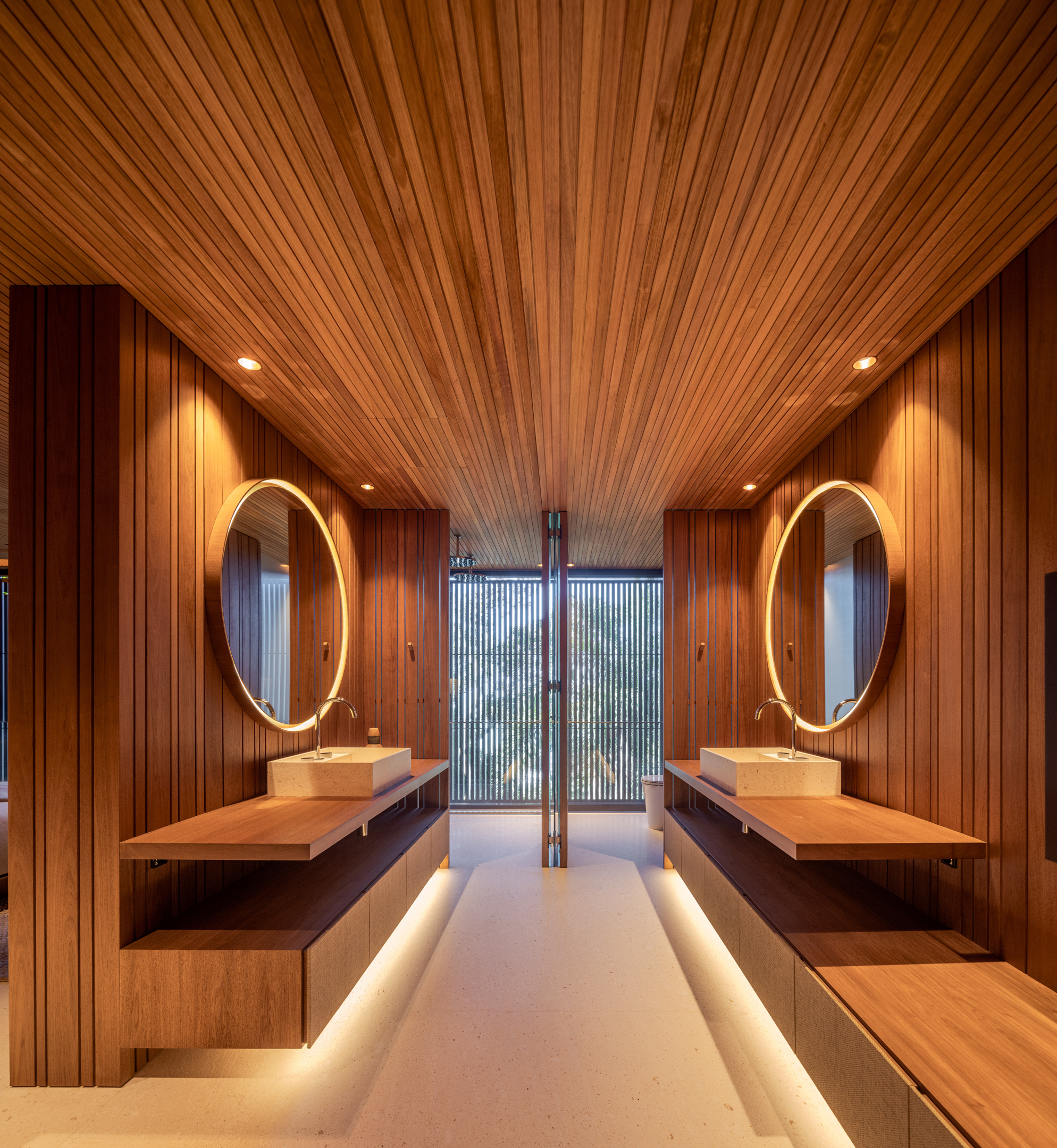 Modern Passive House bathroom with wood-paneled walls and ceiling, two round mirrors above sinks, floating wooden vanities, and a floor-to-ceiling window with blinds.