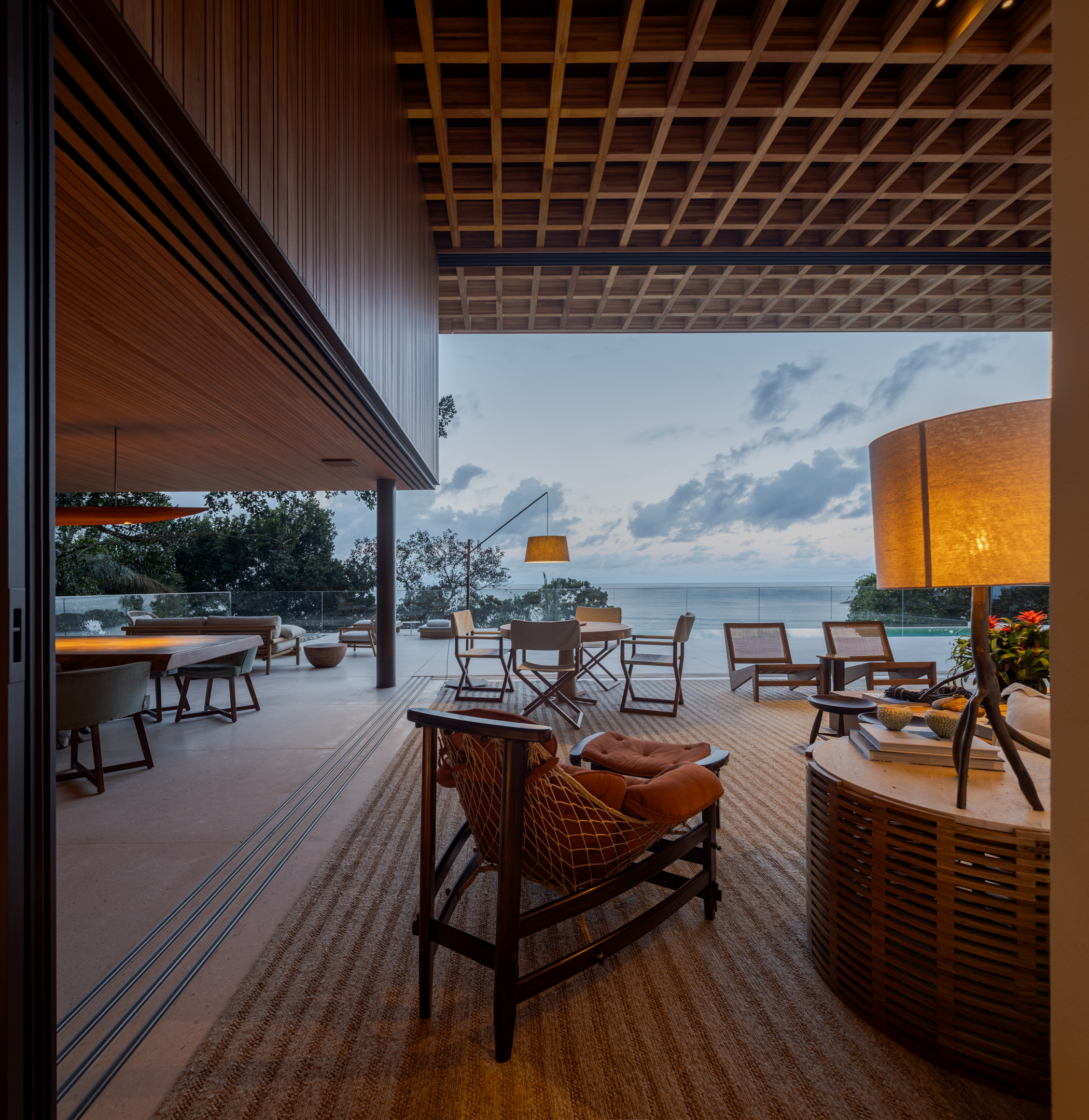 Open-air living room with wooden furniture and lamps, designed with Passive House principles, leading to a terrace with dining tables and chairs overlooking the ocean at sunset.