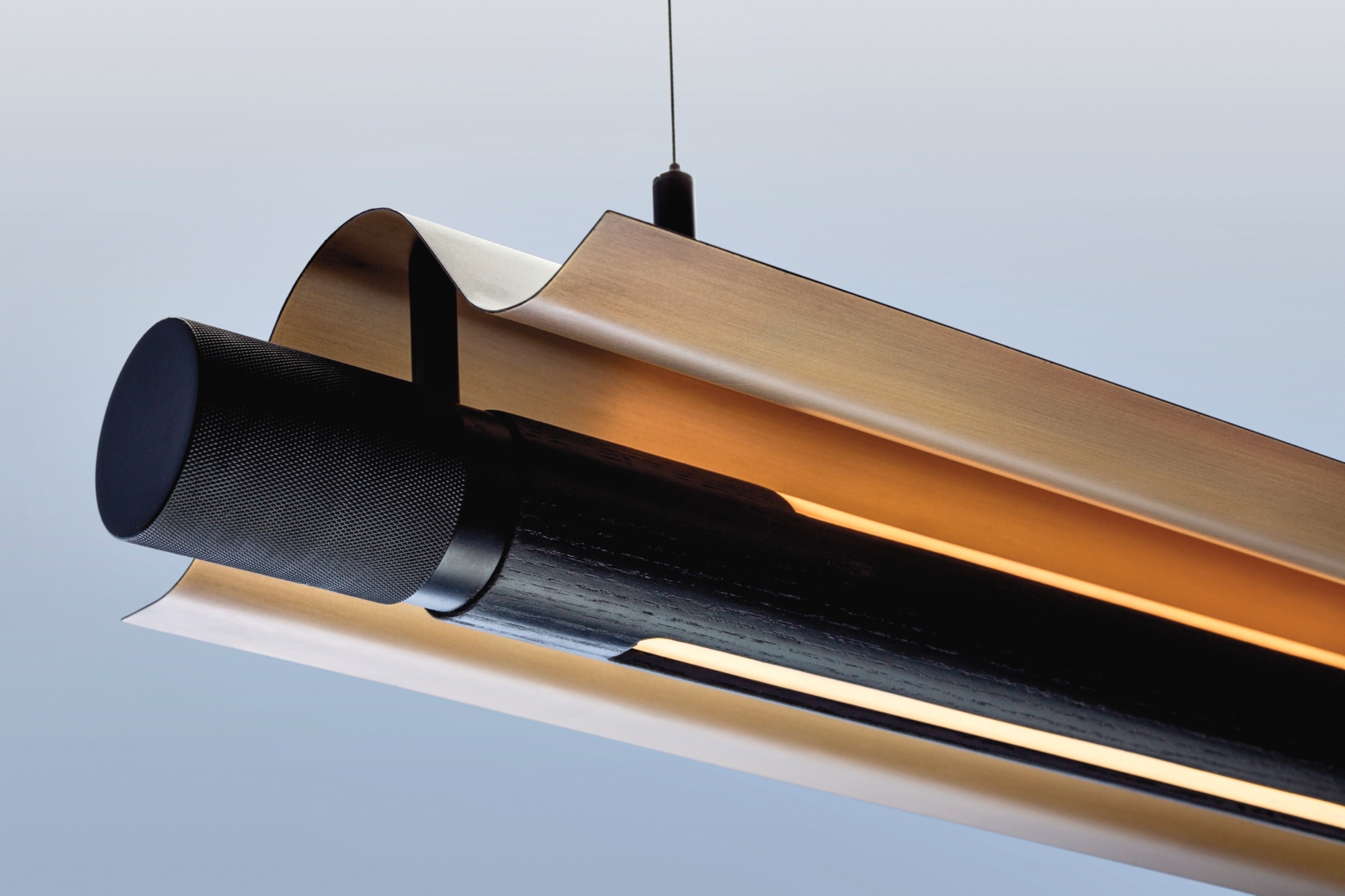Close-up of a modern pendant light fixture with a cylindrical black element and curved metallic shade, suspended by a thin wire against a neutral background.
