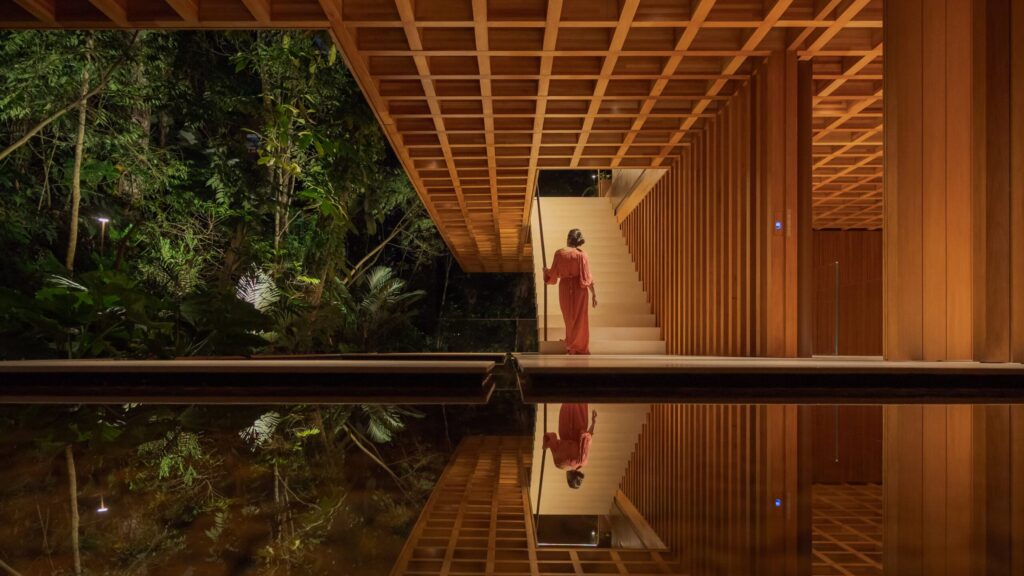 A person in a red dress stands at the base of a staircase in a modern wooden building, reminiscent of the elegant design found in Brazil House, with trees visible outside and their reflection shimmering in a pool of water below.