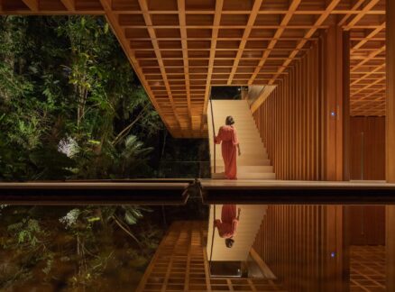A person in a red dress stands at the base of a staircase in a modern wooden building, reminiscent of the elegant design found in Brazil House, with trees visible outside and their reflection shimmering in a pool of water below.