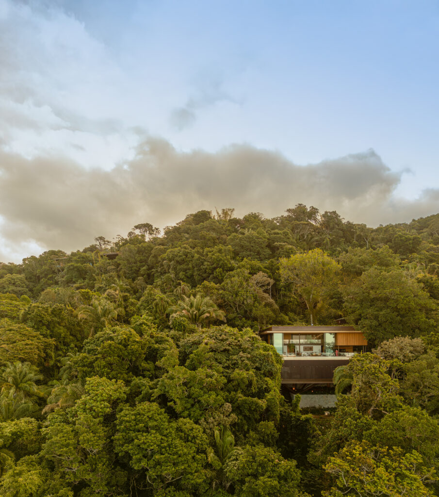 A modern Passive House with large windows is nestled among dense, green trees on a hillside under a partly cloudy sky.