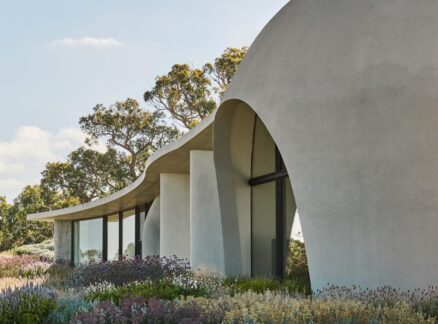 Curved, modern building by Leeton Pointon Architects with large glass windows set among wildflowers and trees under a partly cloudy sky.