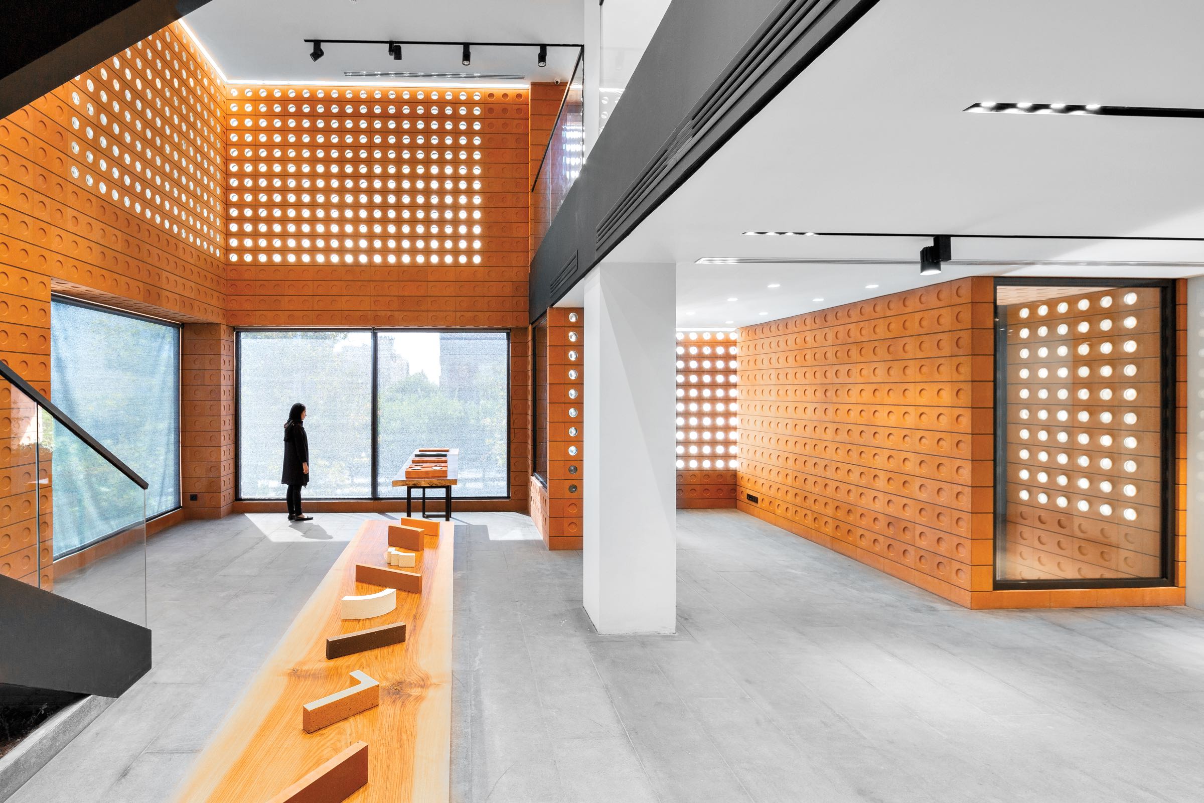 A person stands in a modern, minimalist room by HOOBA Design Group with orange perforated walls, large windows, and sleek wooden furniture.