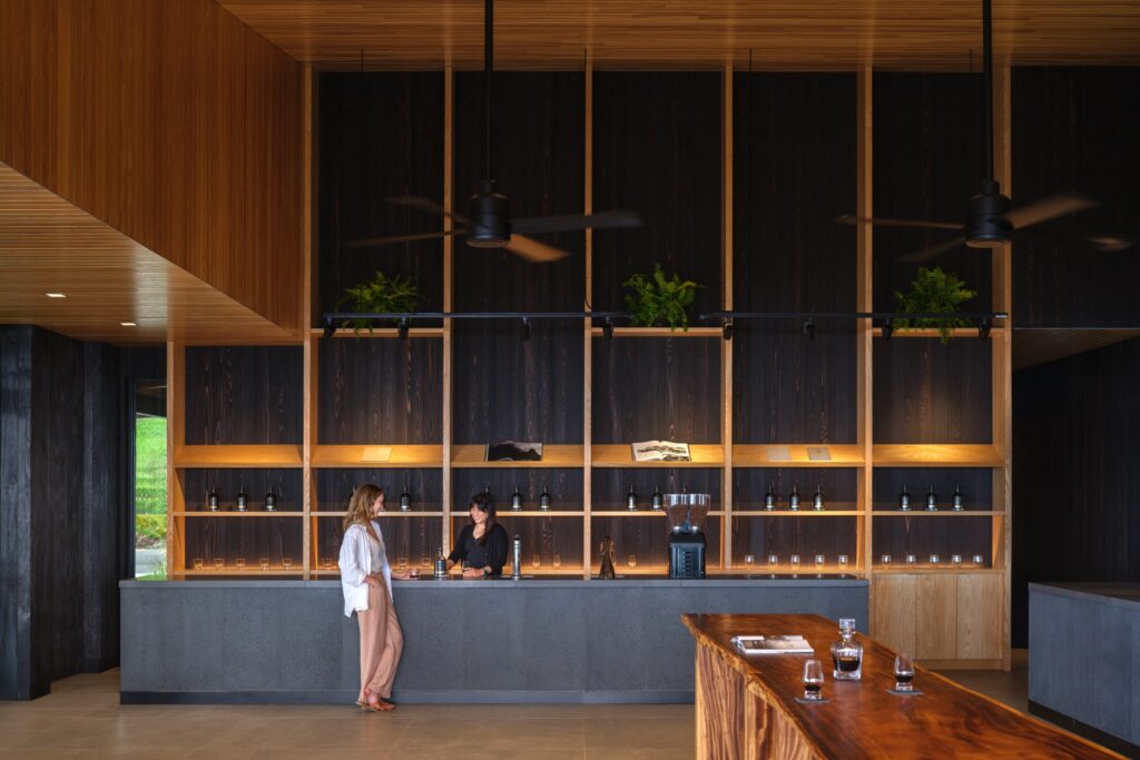 Two people stand at a modern reception desk with wooden shelves behind them, containing plants, bottles, and decorative items in a spacious, well-lit lobby.