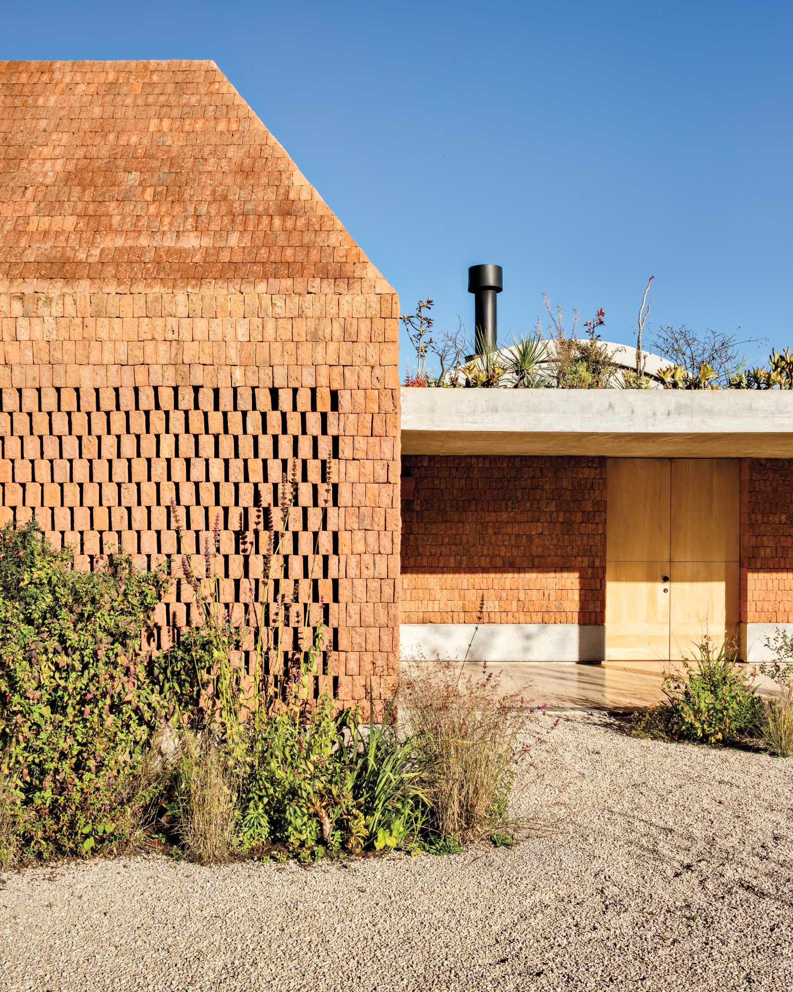 Built by Fernanda Canales, a modern house with textured brick walls, a wooden door, rooftop greenery, and surrounding gravel and plants under a clear blue sky.