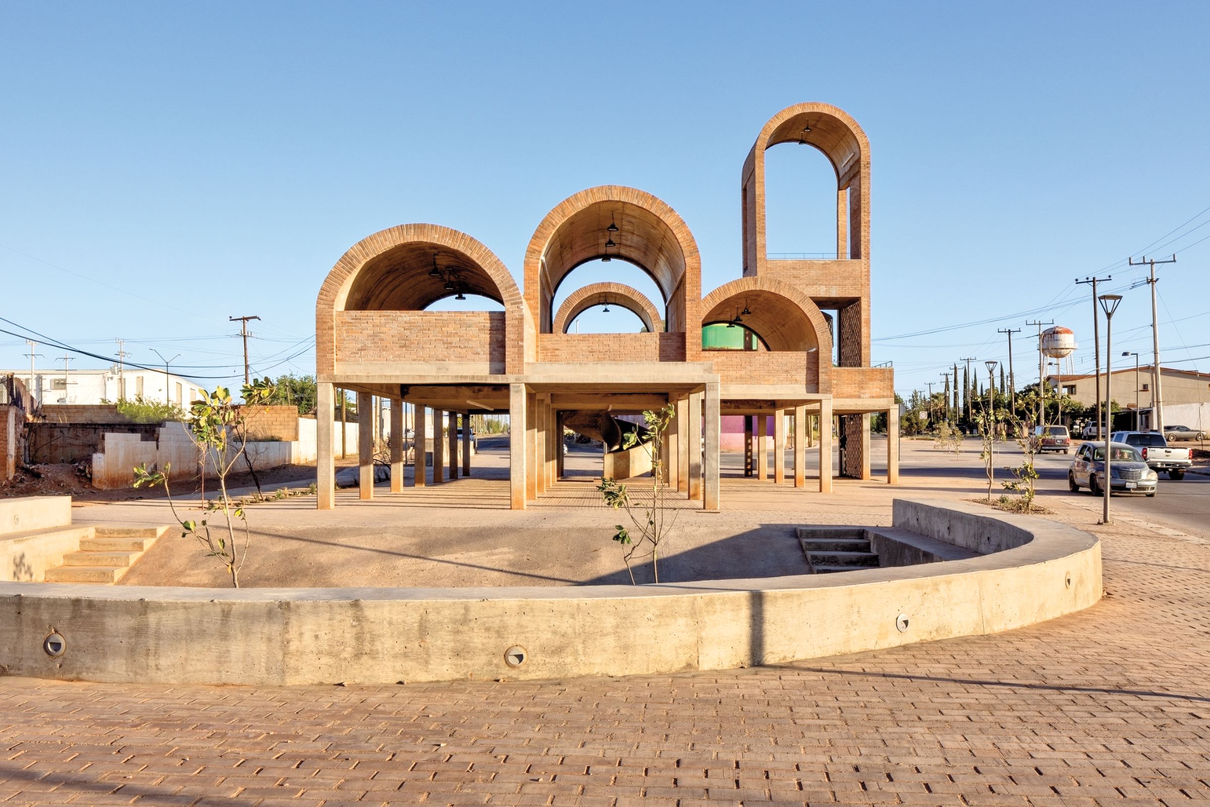 A modern brick structure by Fernanda Canales with large arches and open walls stands in a paved plaza under a clear blue sky, surrounded by a few small plants and nearby roads.