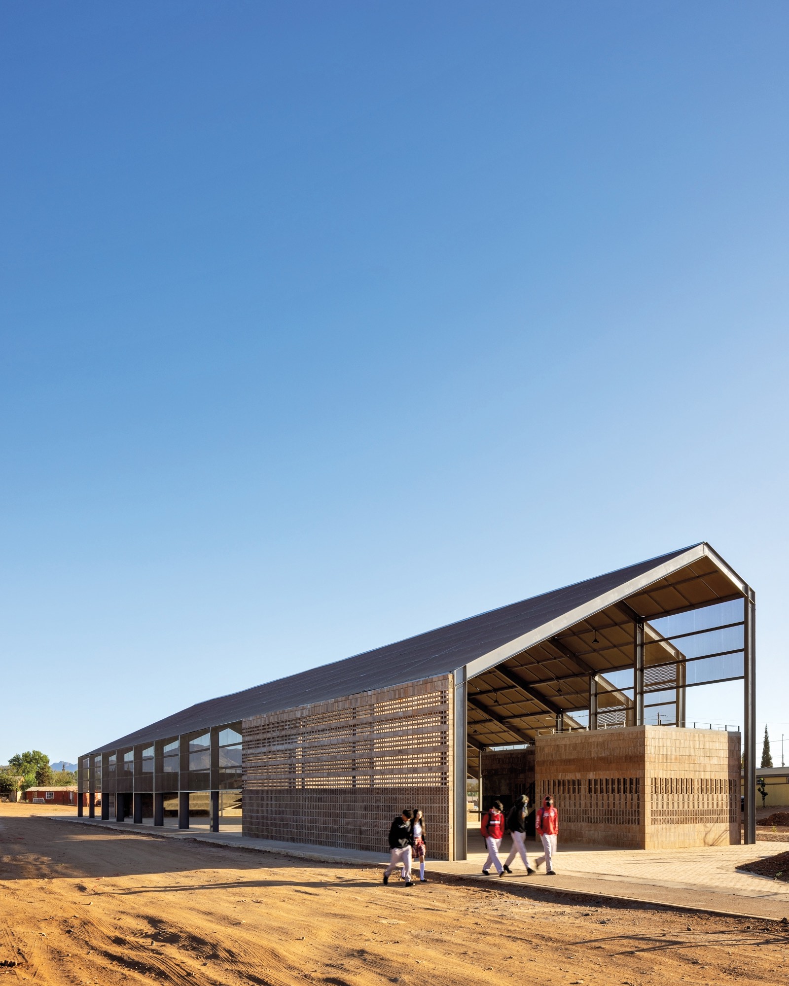 A modern open-sided building with a sloped metal roof sits on sandy ground under a clear blue sky. Four people are walking near the entrance.