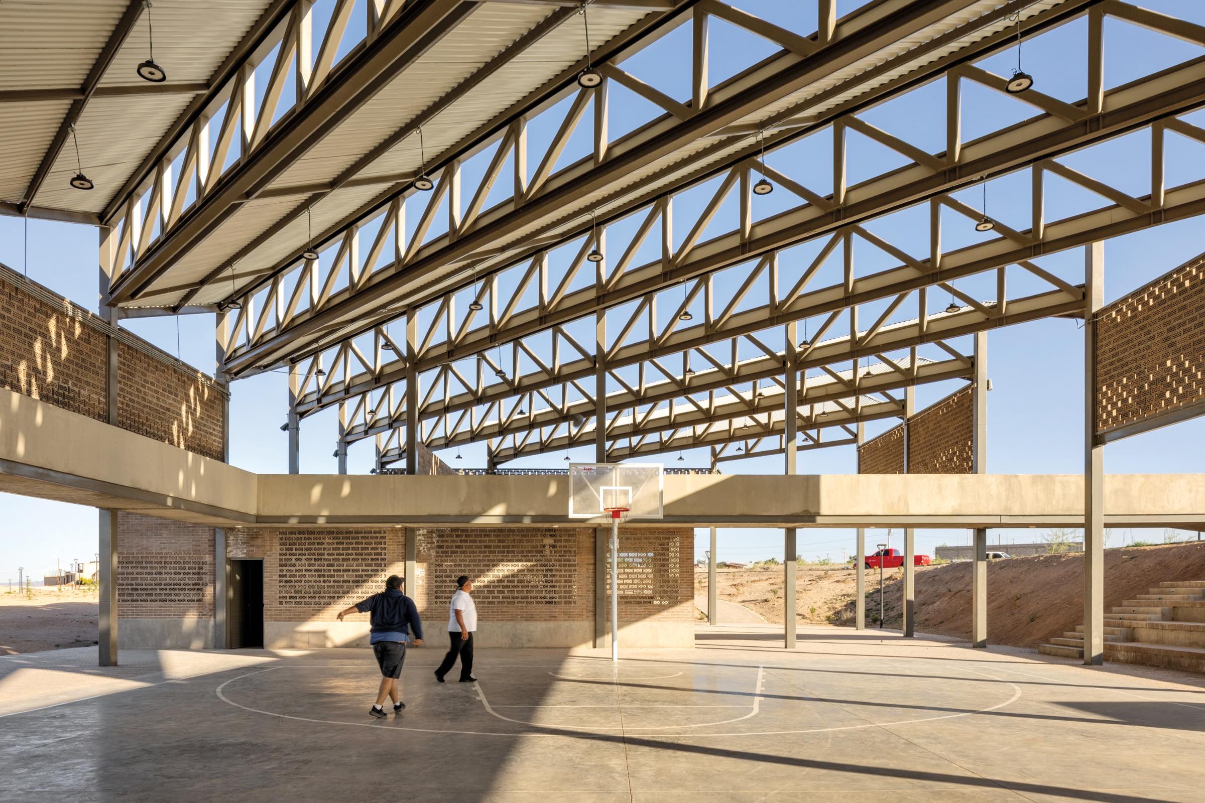 Two people walk on the concrete floor of an open-air basketball court inside a large, modern metal and brick structure with exposed beams and natural light.