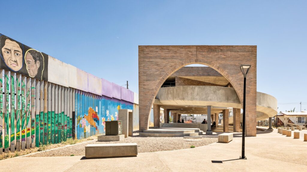 Outdoor plaza by Fernanda Canales with a modern concrete and brick pavilion, curved archway, benches, and a mural-covered border wall featuring painted faces and landscapes under a clear blue sky.