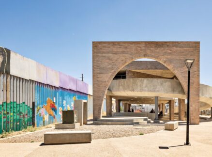 Outdoor plaza by Fernanda Canales with a modern concrete and brick pavilion, curved archway, benches, and a mural-covered border wall featuring painted faces and landscapes under a clear blue sky.