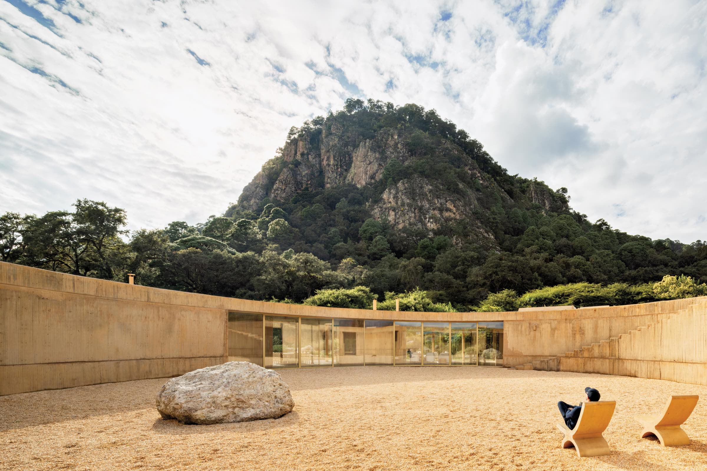 CASA 720 by Fernanda Canales: Concrete-walled courtyard with a large rock, glass-walled building, lounge chairs, and a person sitting; forested mountain rises in the background under a partly cloudy sky.