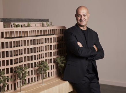 A man in a black suit stands with arms crossed next to a detailed architectural model of a multi-story building with trees.