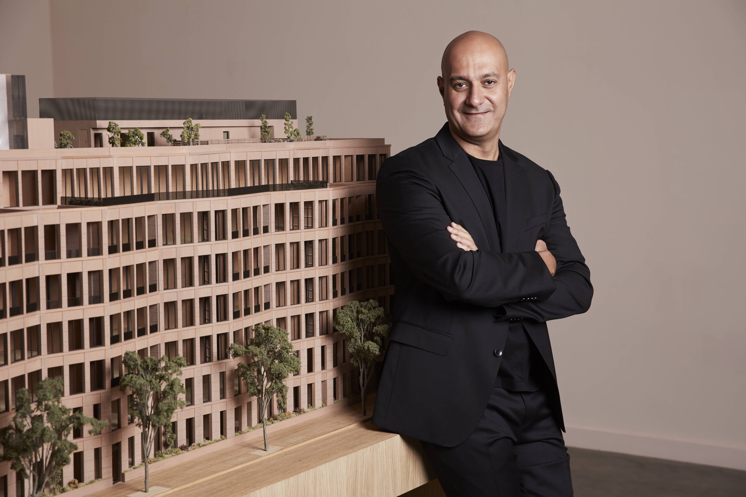 A man in a black suit stands with arms crossed next to a detailed architectural model of a multi-story building with trees.