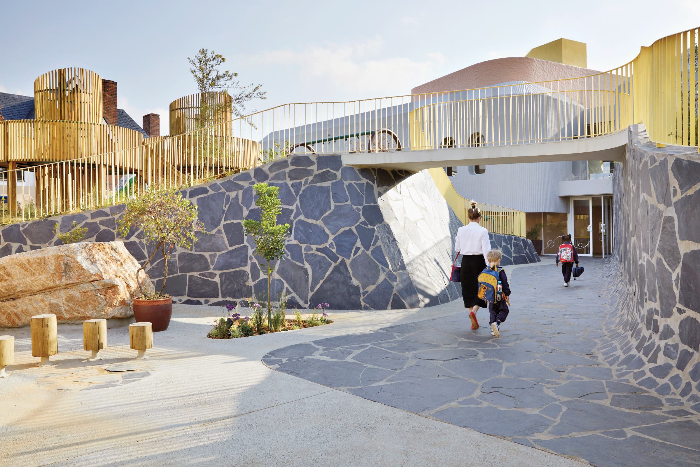 Hubo Studio: A woman and two children walk on a stone path toward a modern building with geometric shapes and yellow railings, under a bridge.