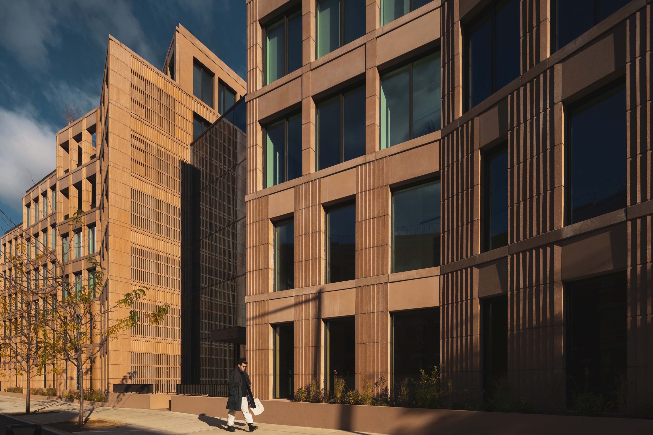 Person walks on a sidewalk beside a modern, tan-colored building with large windows and geometric architectural features under sunlight.