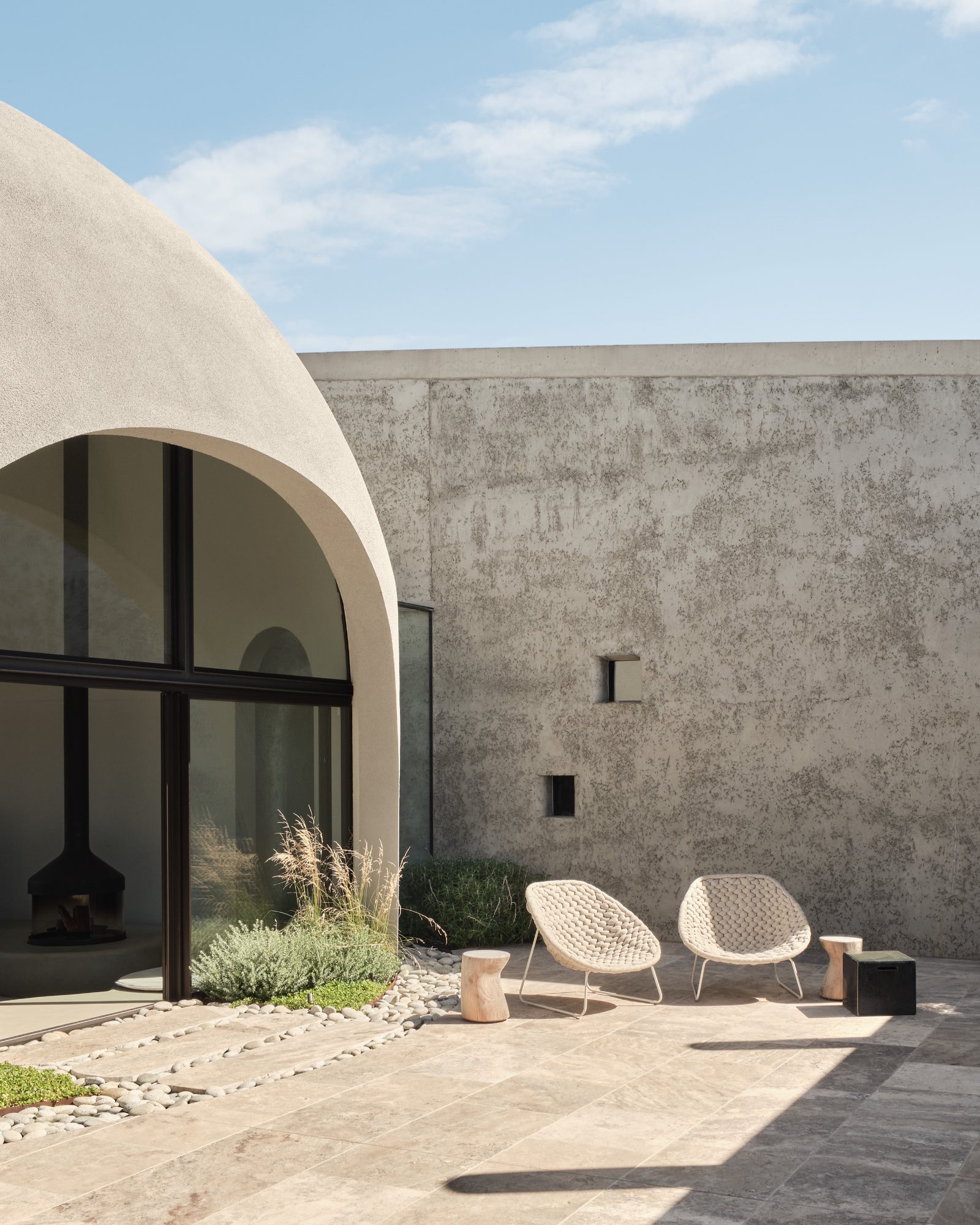 Modern patio by Leeton Pointon Architects with two woven chairs, small tables, and minimalist landscaping beside an arched window and textured concrete wall under a clear sky.