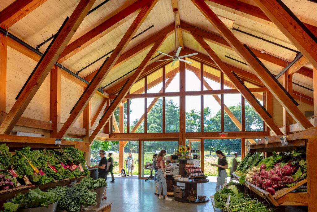Interior of a wooden market building with exposed beams, fresh produce on display, and several people shopping on a sunny day.