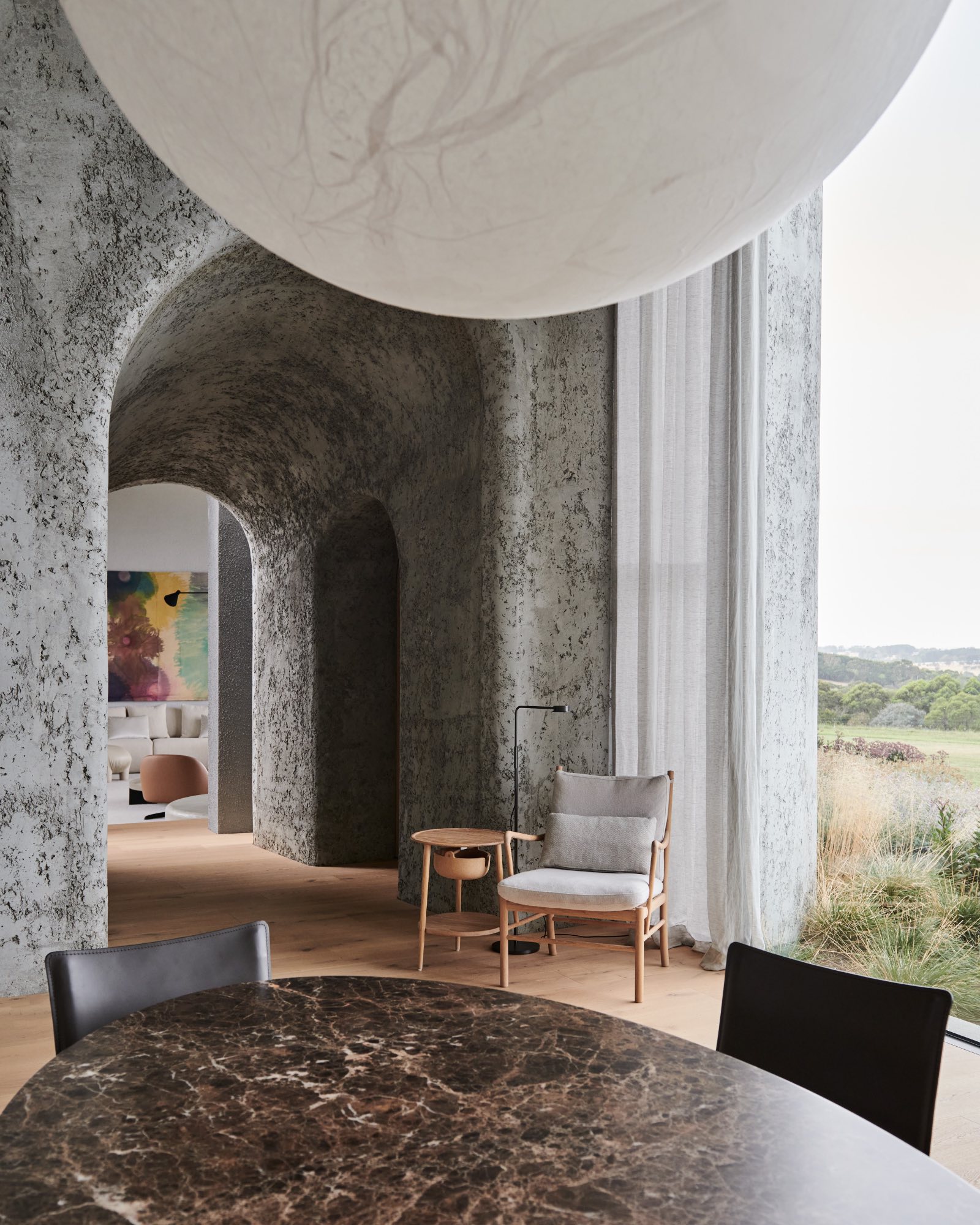 Modern interior by Leeton Pointon Architects with textured gray archways, a round brown marble table, black chairs, a wooden lounge chair, and large windows overlooking a natural landscape.