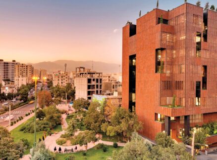 HOOBA Design Group Crafts with Materials and Light A modern, multi-story building with a reddish-brown facade stands beside a green urban park at sunset, with city buildings and distant mountains in the background.