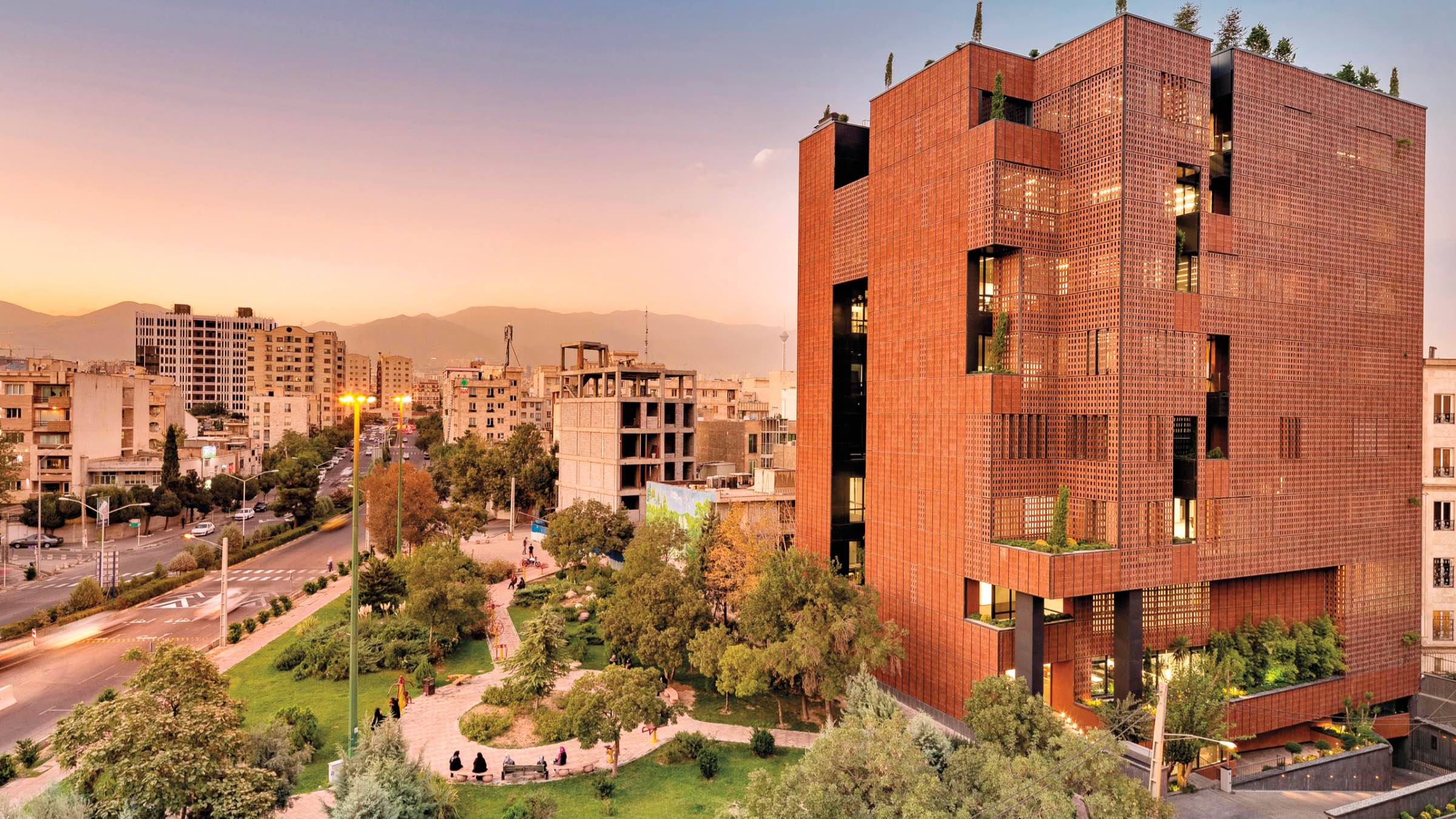A modern, multi-story building by HOOBA Design Group with a reddish-brown facade stands beside a green urban park at sunset, with city buildings and distant mountains in the background.