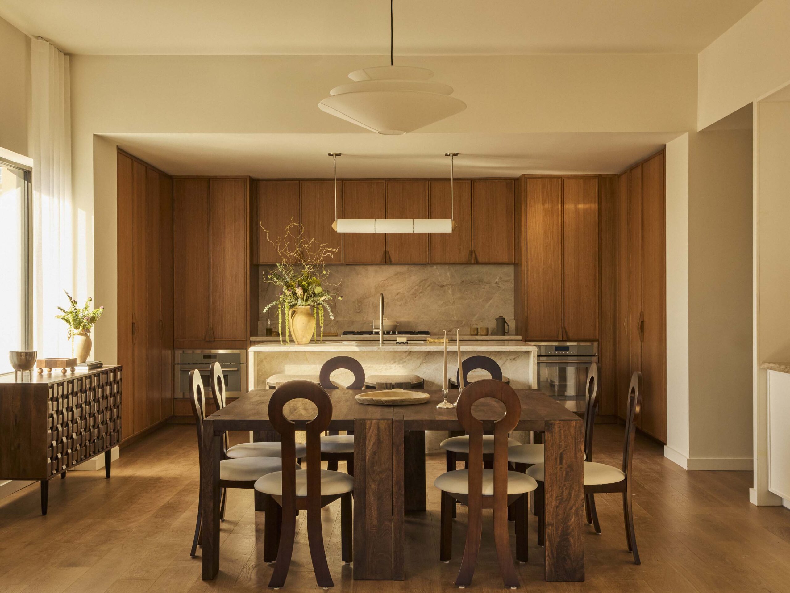 Modern kitchen and dining area with wooden cabinets, a dark wood dining table, six chairs, marble backsplash, minimalist decor, and natural light from a large window.