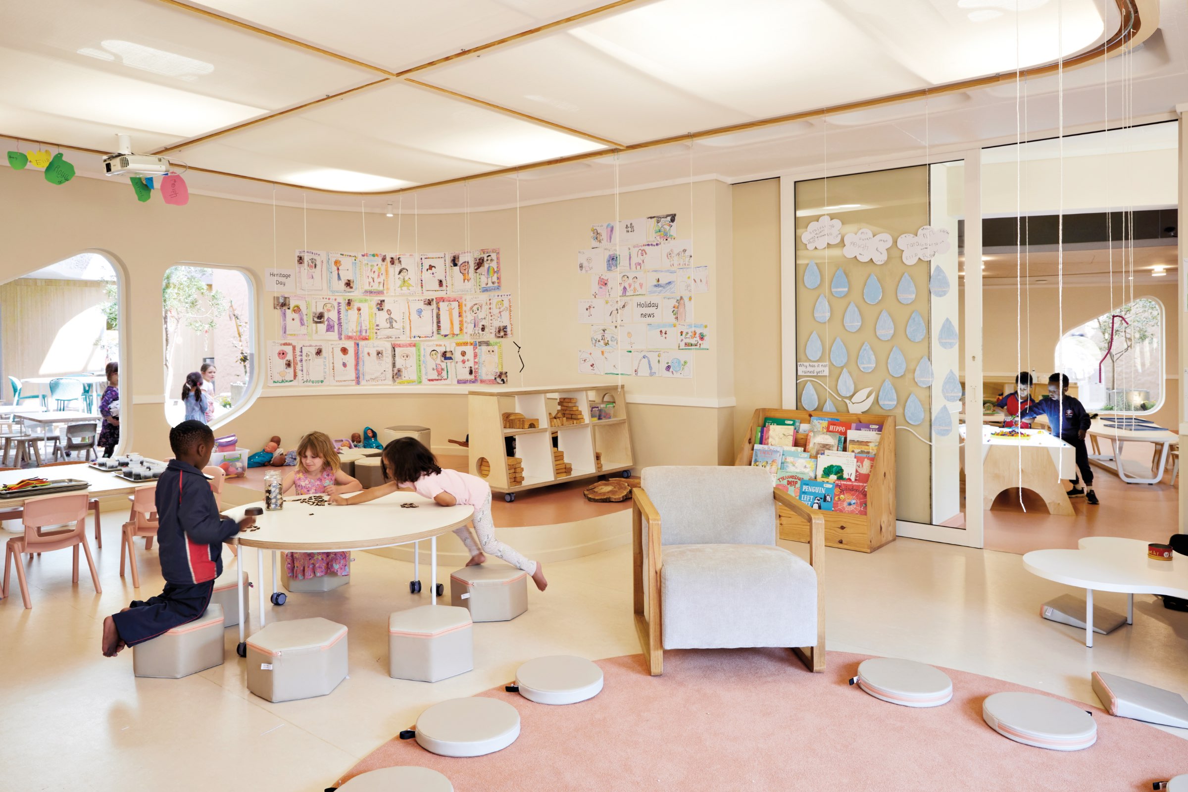 Children are playing and drawing in a brightly lit classroom by Hubo Studio decorated with art and books, featuring various seating areas and educational displays.