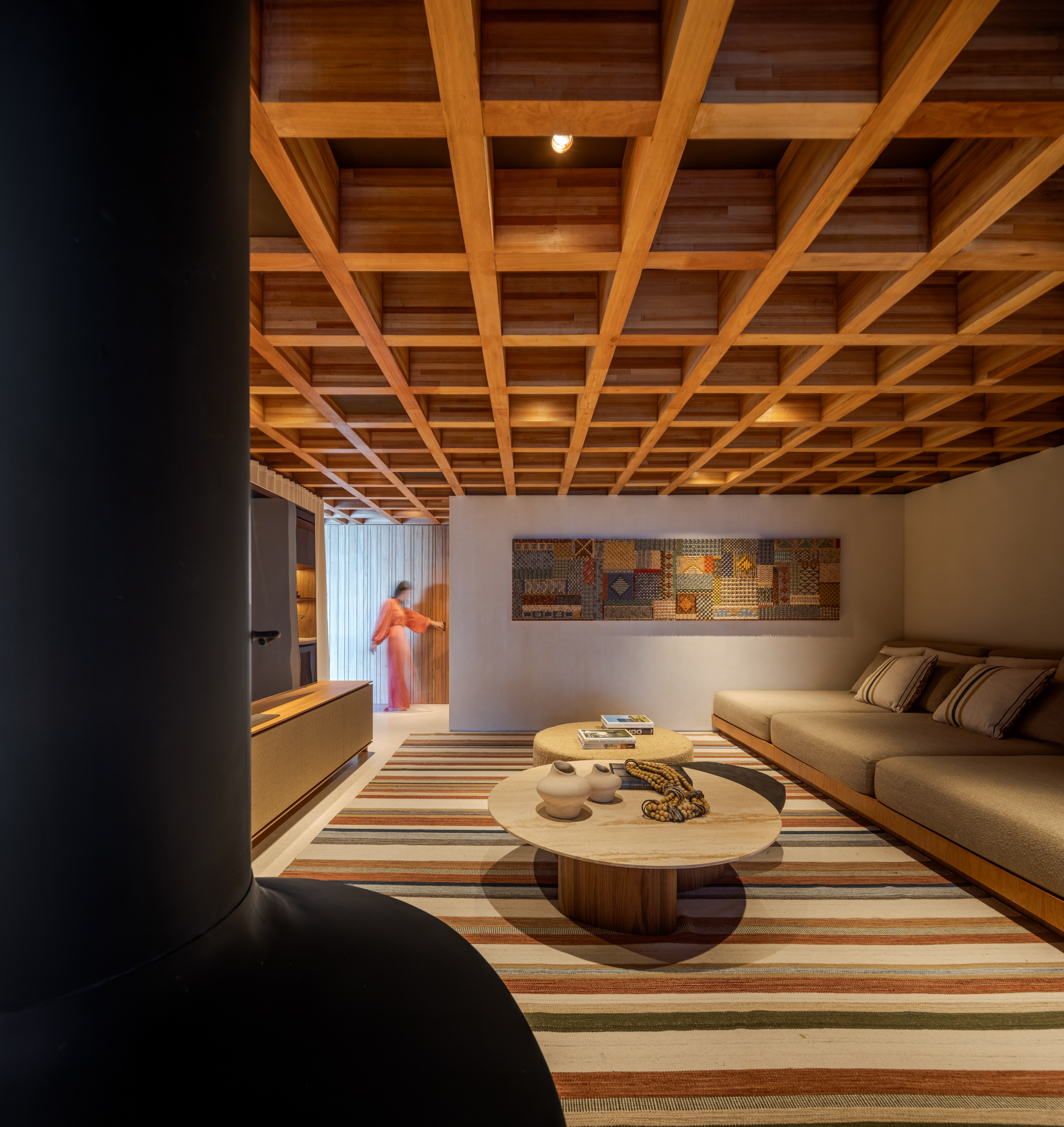 A modern Passive House living room with a wooden grid ceiling, striped rug, low table, beige sofa, wall art, and a person in the background near a doorway.