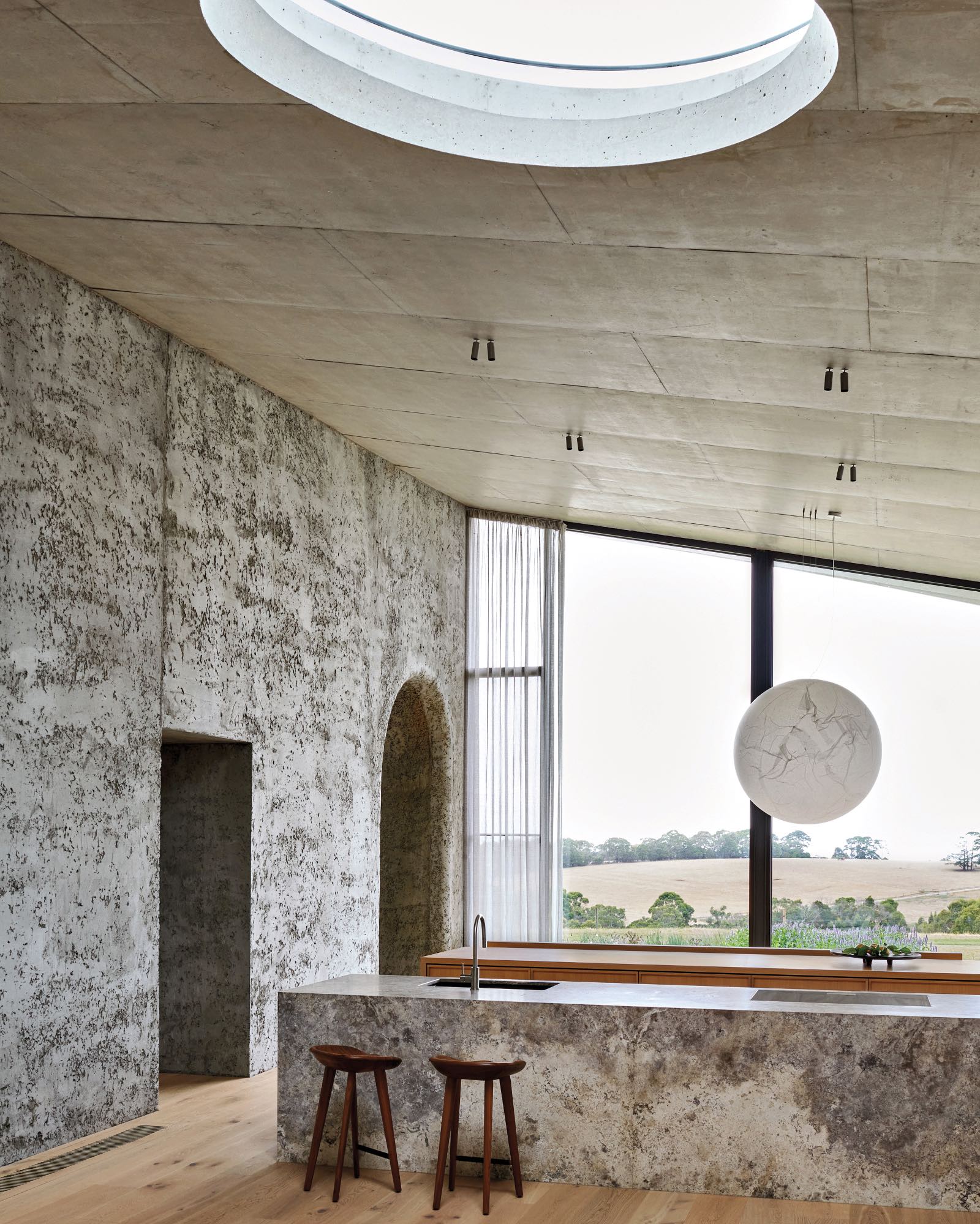 Modern kitchen with textured concrete walls, a stone island with a sink and two wooden stools, large window with countryside view, round ceiling skylight, and pendant light.