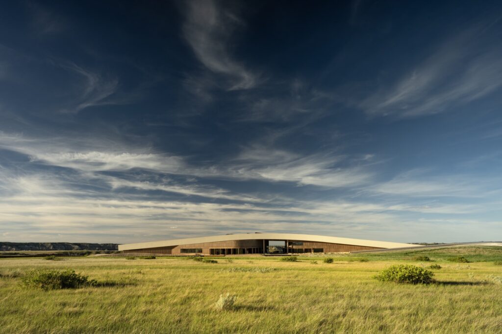 A modern, low-profile building with a flat roof sits in a grassy, open landscape under a dramatic sky with wispy clouds.