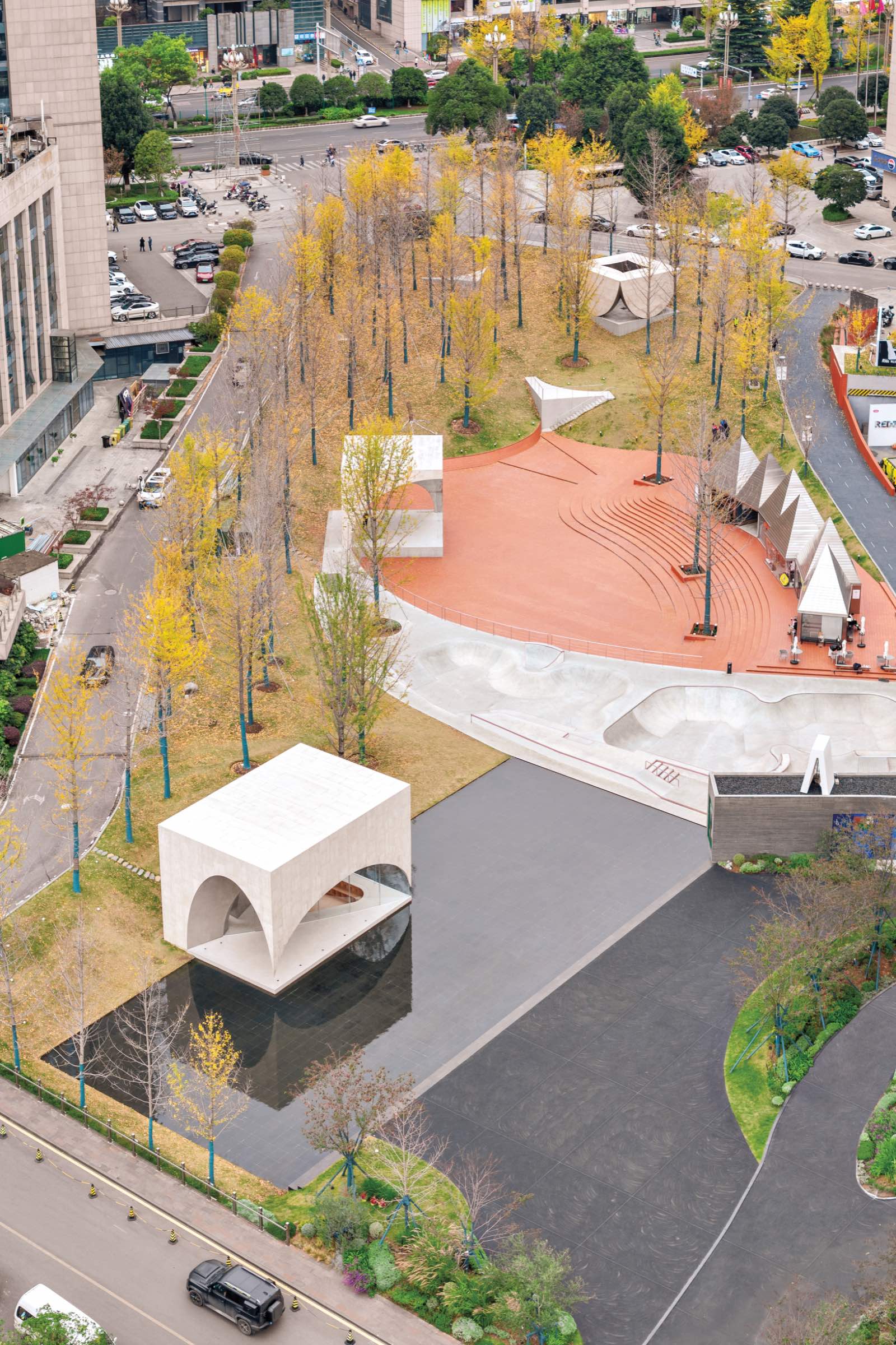 Aerial view of a city park by Atelier Xi with geometric white structures, a red circular plaza, walking paths, trees with yellow leaves, and adjacent roads with cars.