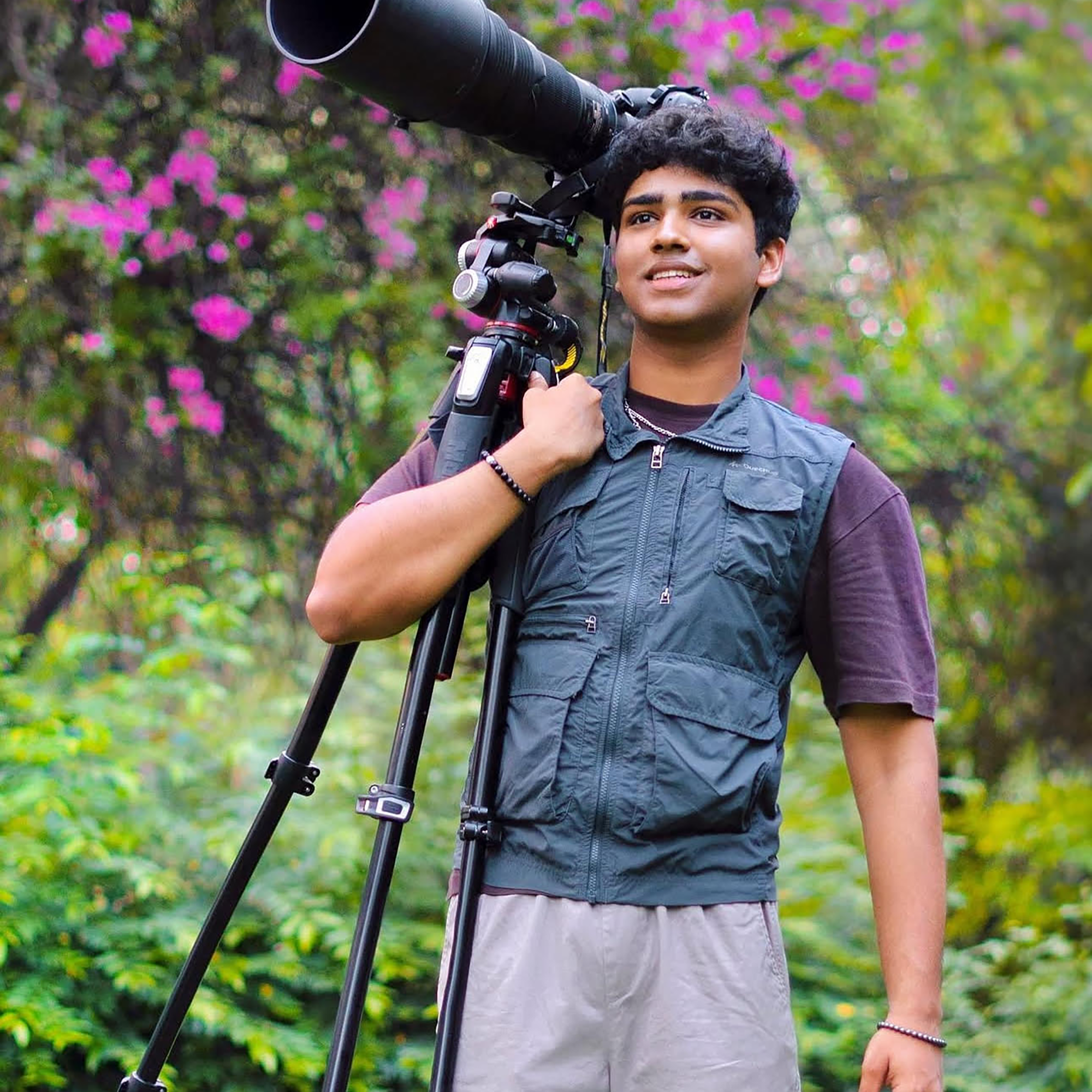 A man outdoors holds a large camera with a telephoto lens mounted on a tripod, surrounded by greenery and pink flowers, capturing the inspiring moments of the Trailblazer Awards.