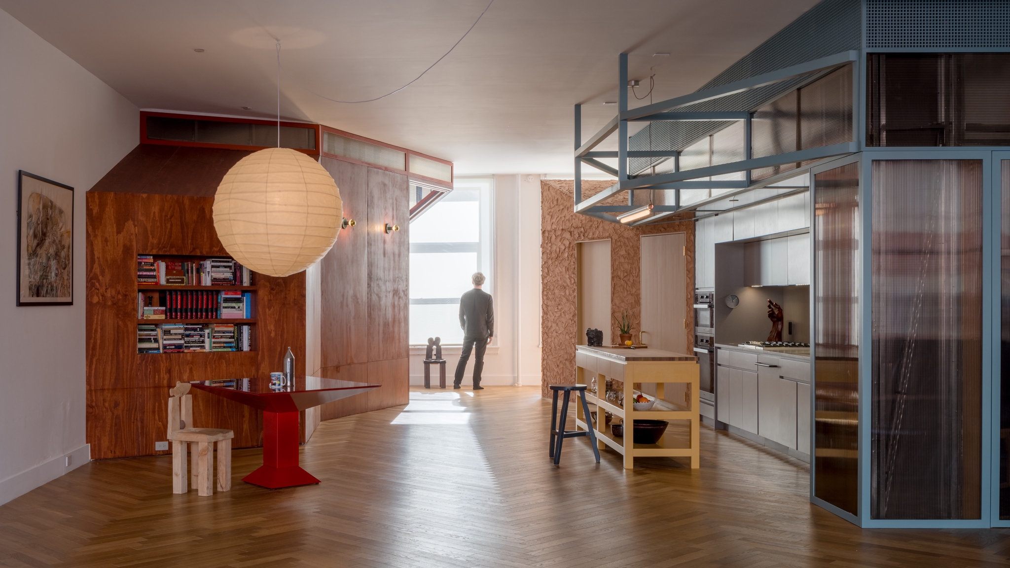 Modern apartment interior with wooden floors, a dining area, open kitchen, thoughtfully arranged shelving, and a person standing by a large window looking outside—all reflecting principles of Antifascist Architecture through open spaces and welcoming design.