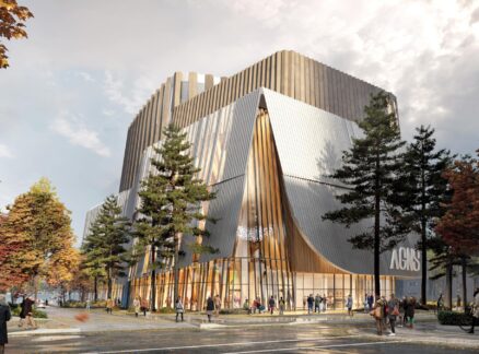 Modern multi-story cultural building by Omar Gandhi with tall windows, unique angular design, and people walking nearby. Surrounded by trees with autumn foliage under a cloudy sky.
