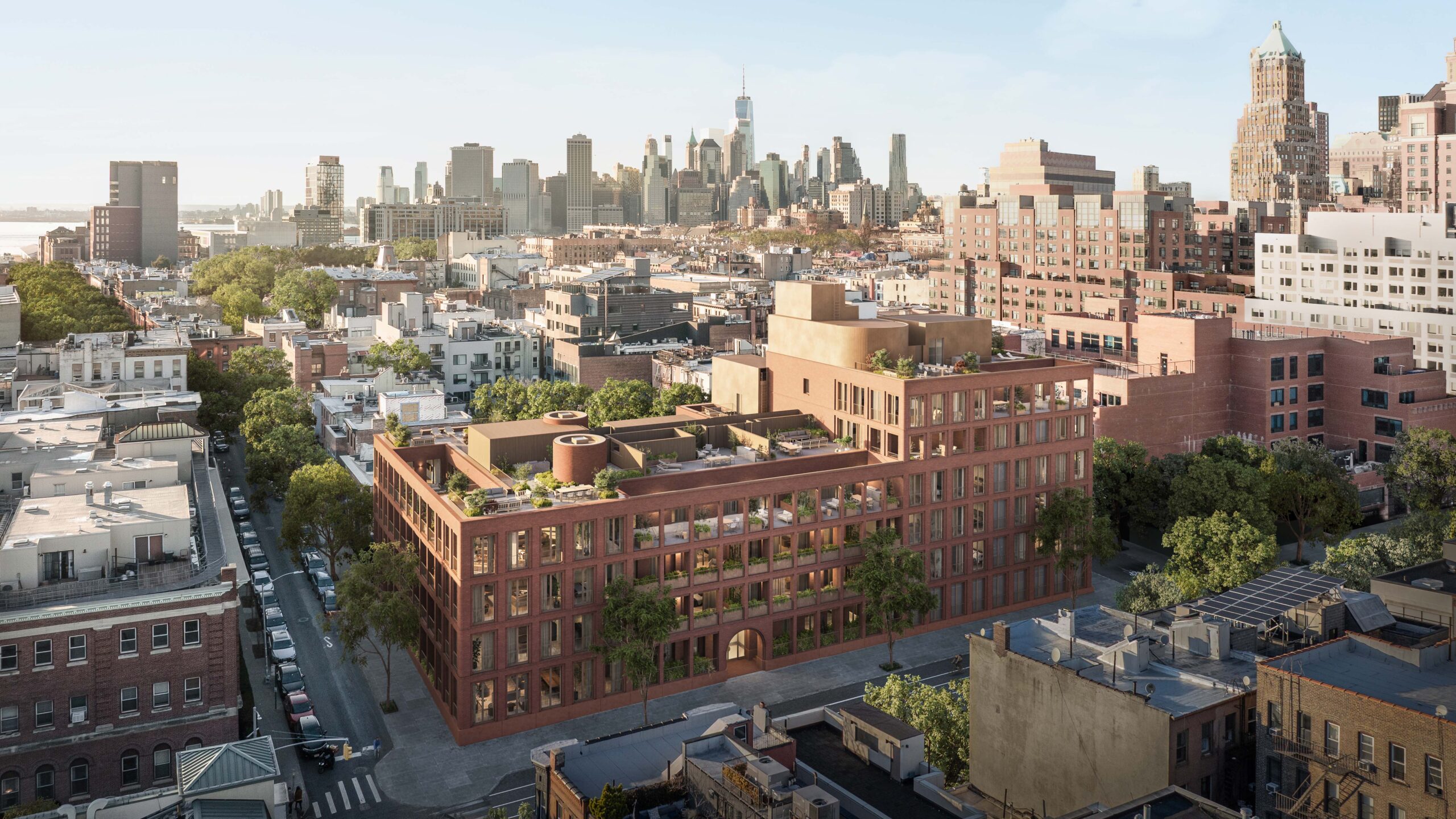 A brick midrise building with rooftop terraces stands among city blocks, with a view of a downtown skyline in the background under clear skies.