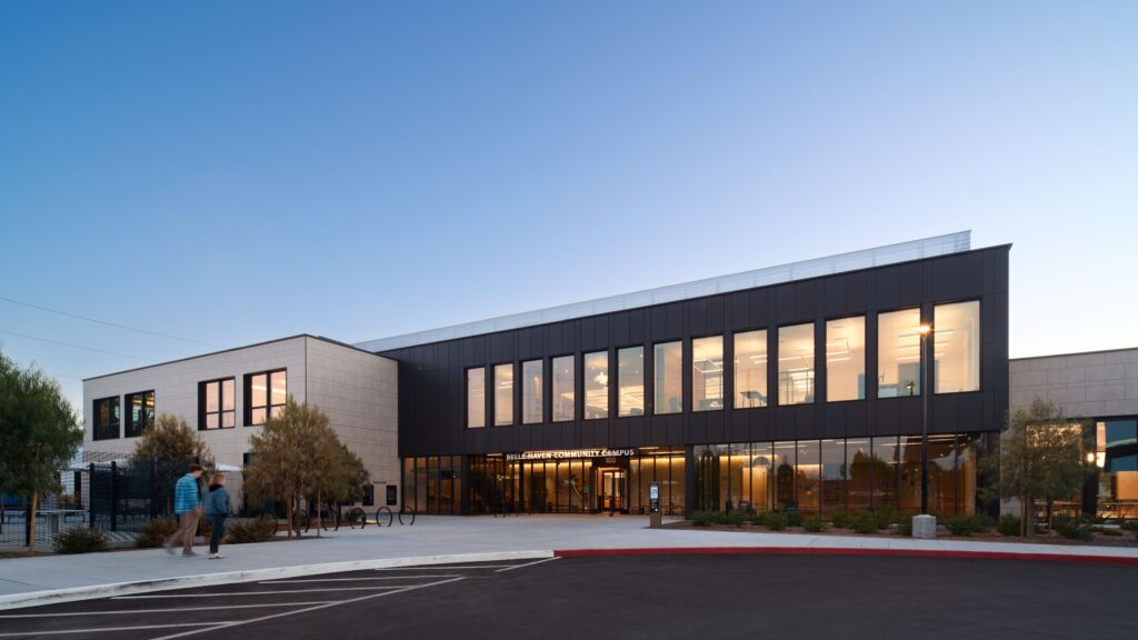 Modern two-story building with large windows at dusk, lights on inside, a few people walking near the entrance, and a parking lot in the foreground.