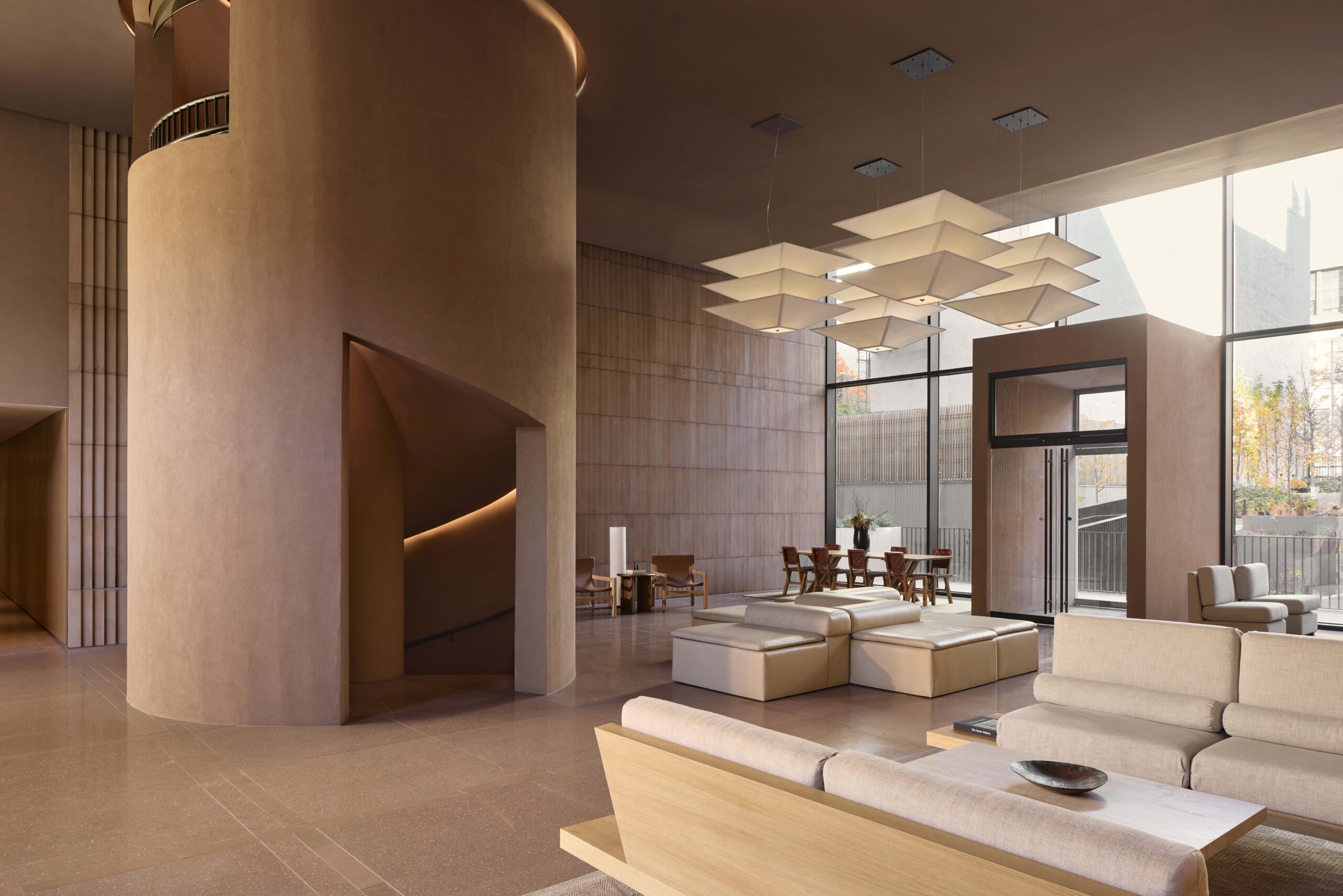 Modern lobby with beige furnishings, geometric light fixtures, large windows, and a curved stairwell. Tables and chairs are arranged near the windows, allowing natural light to fill the space.