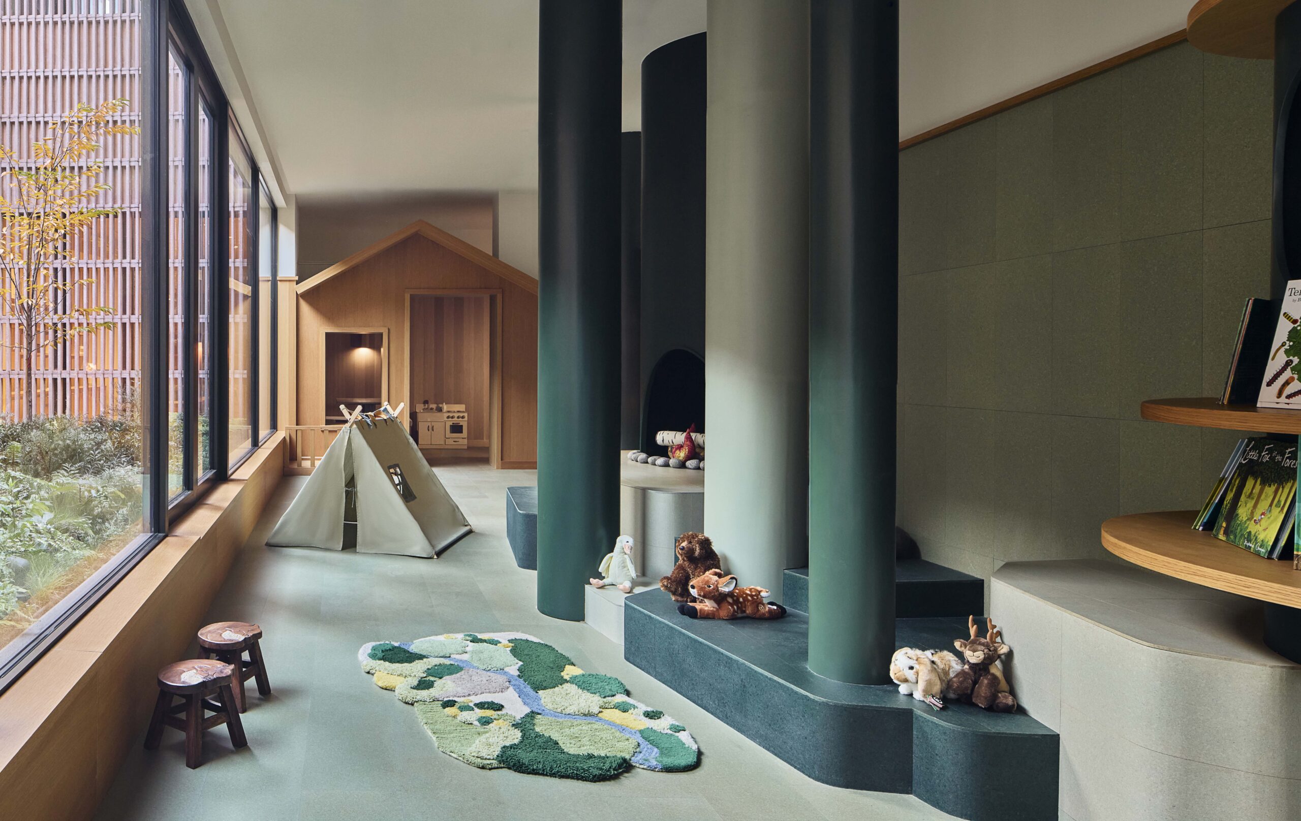 Children’s playroom with green pillars, a tent, a playhouse, animal plush toys, bookshelves, wooden stools, and a window overlooking a garden.