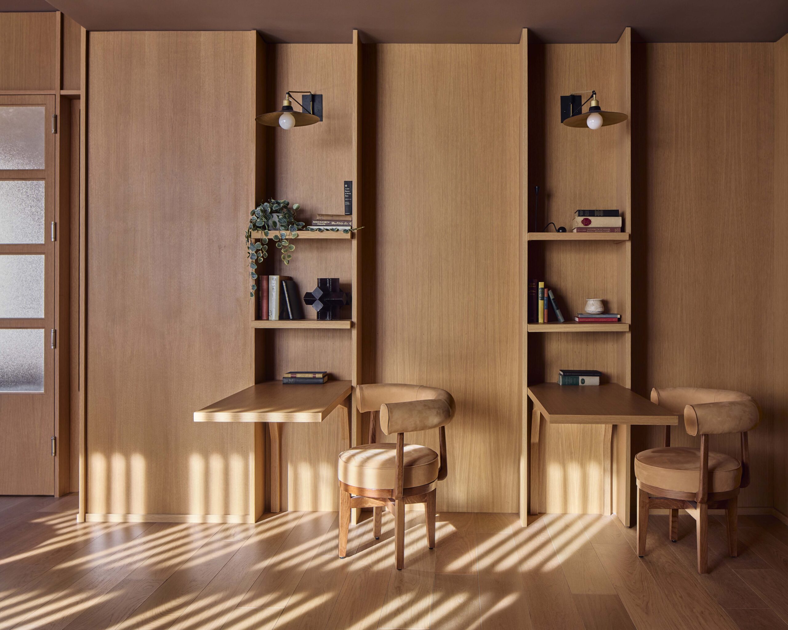 Two built-in wooden desks with shelves and chairs against a wooden wall, each desk lit by a wall sconce, with books, plants, and decorative items on the shelves. Light streams in from the left.