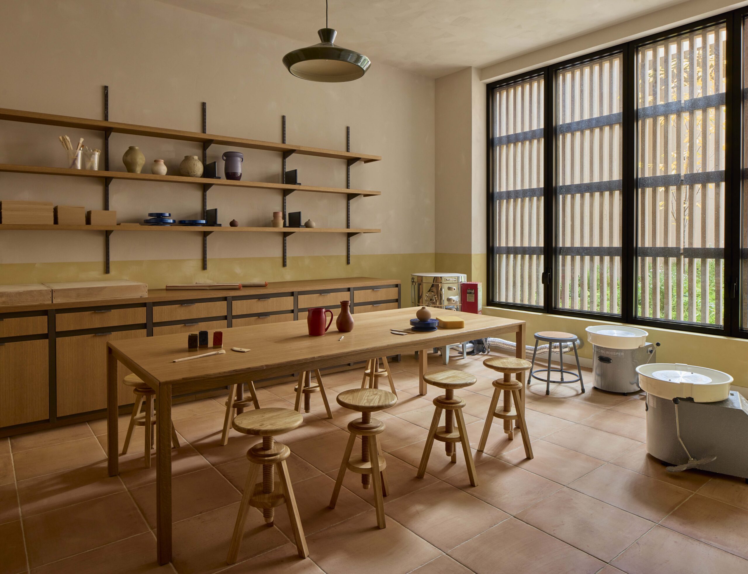 A pottery studio with a long wooden table, several stools, open shelves holding pottery and supplies, large windows, and two pottery wheels.