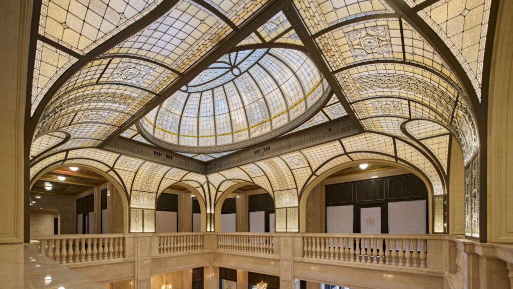 Interior view of a grand hall with a large stained glass dome ceiling, arched windows, and ornate stone railings.