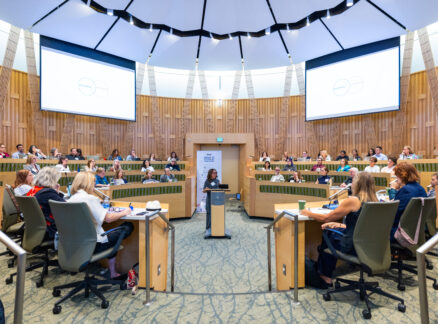 A woman speaks at a podium in the center of a modern, circular lecture hall filled with seated attendees. Two large projection screens are visible on the walls.