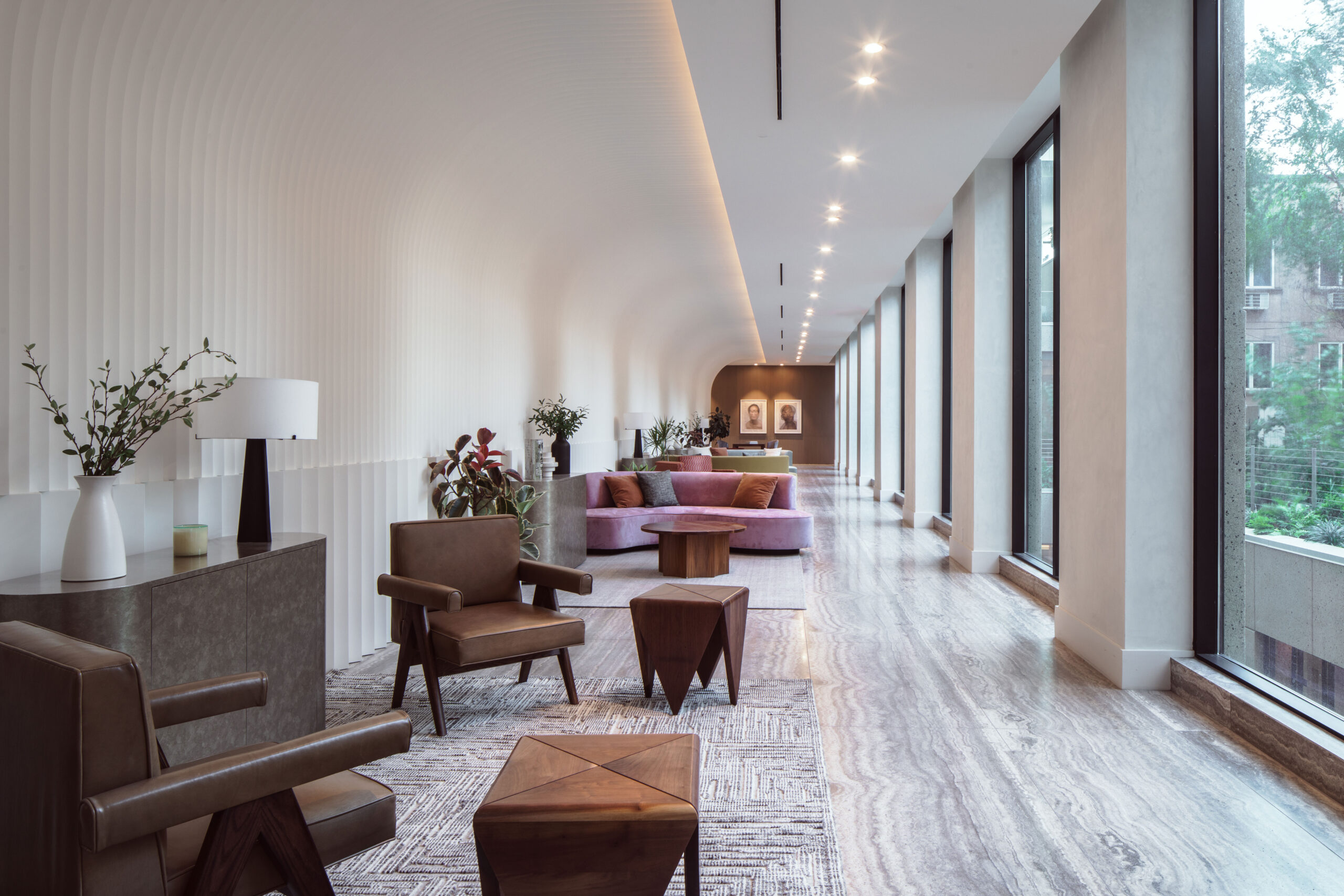 Modern lounge with leather chairs, geometric tables, and a purple sofa along a long hallway with large windows and natural light.