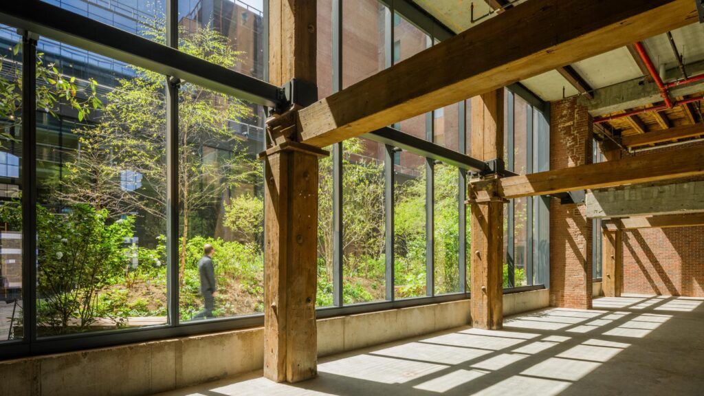Large windows in an industrial-style building reveal a green outdoor garden with a person walking by; sunlight creates shadows on the concrete floor, highlighting the sustainable design that earned recognition from the Planet Positive Awards.