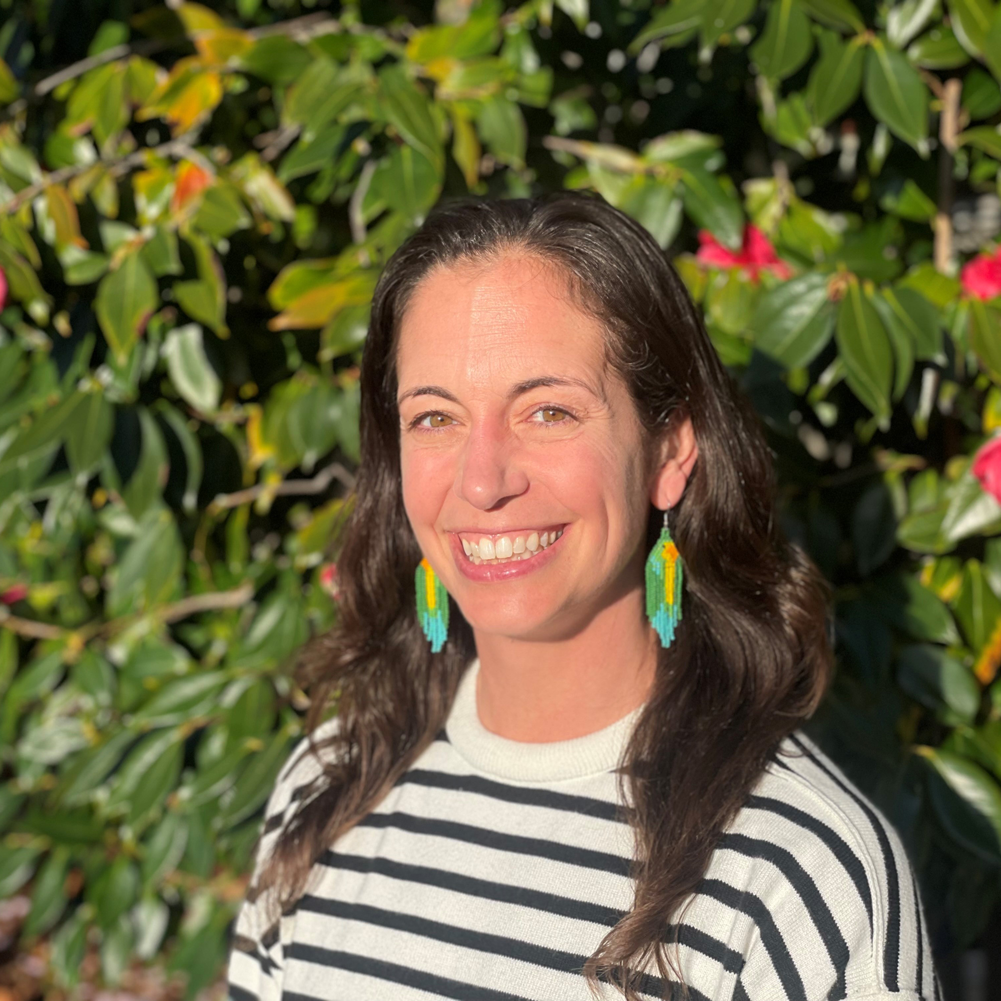 Woman with long brown hair, wearing a striped sweater and colorful earrings, smiles outdoors in sunlight with leafy green plants and pink flowers in the background, radiating the confidence of a Trailblazer Awards honoree.