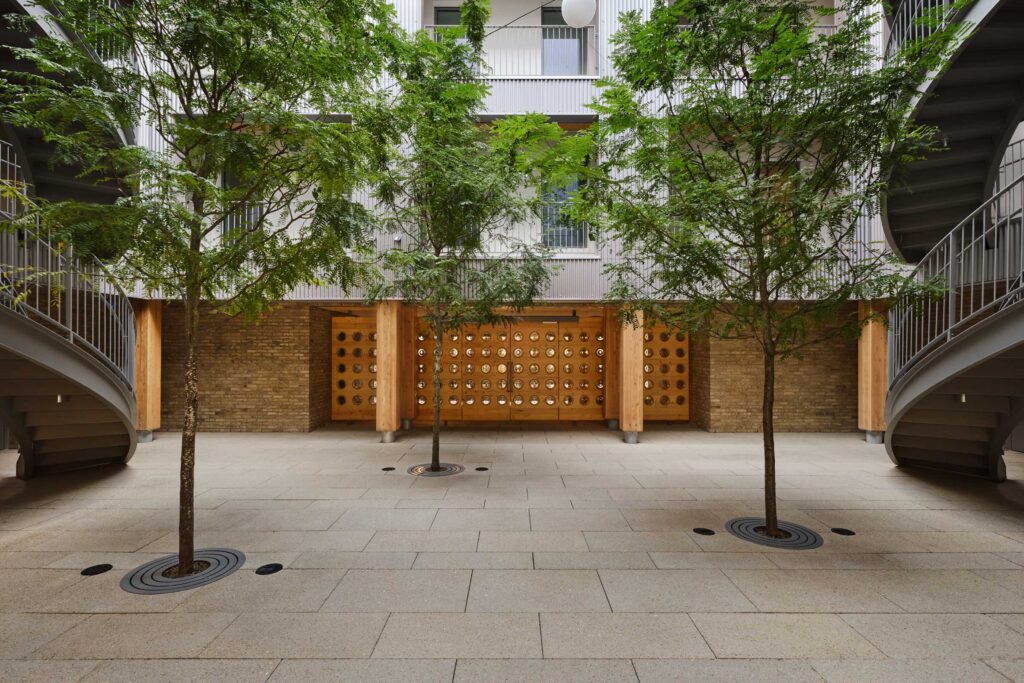 A modern courtyard with three trees, light-colored paving, brick walls, circular staircases on both sides, and a balcony above.