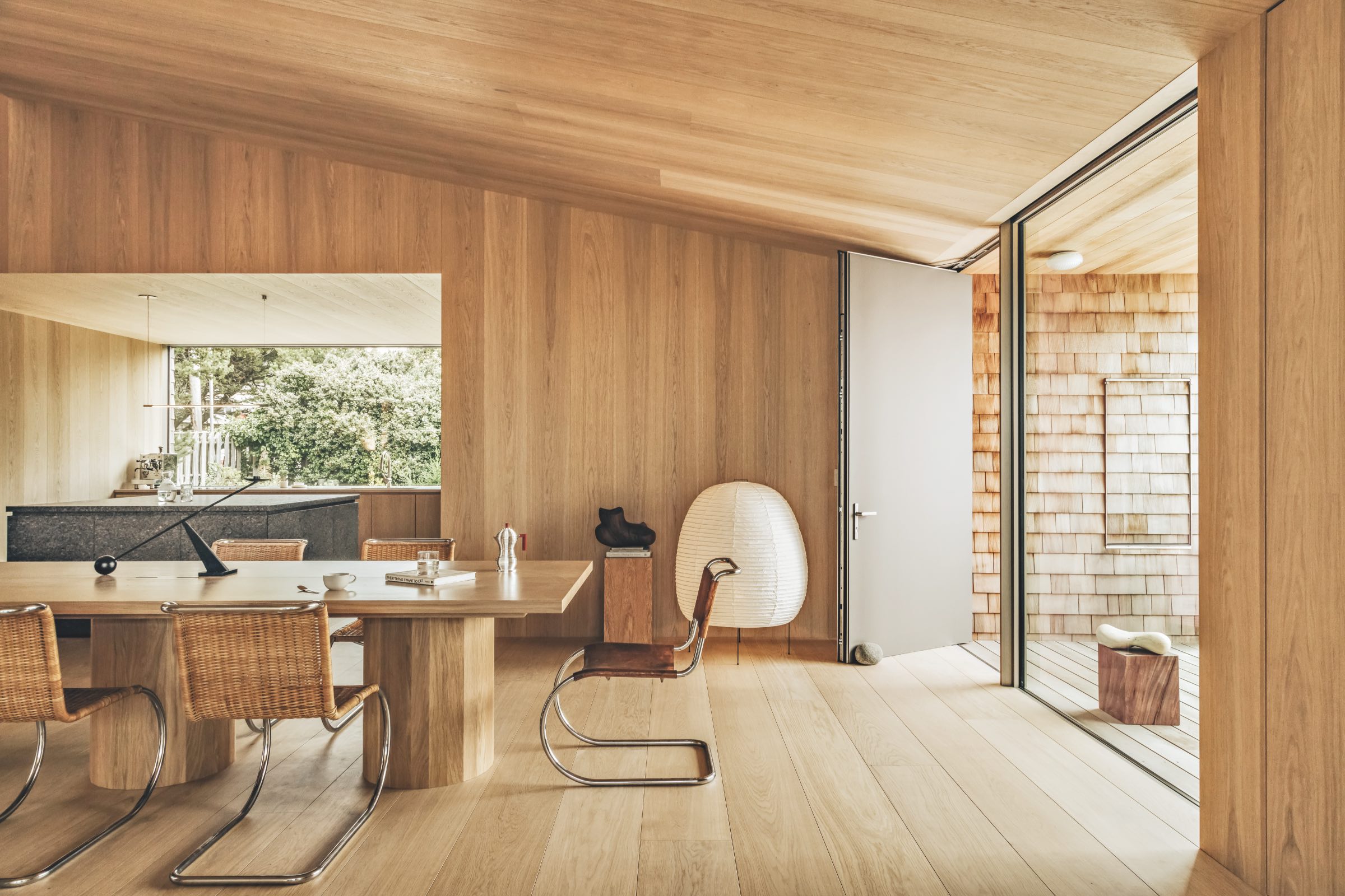 Minimalist dining room with wood-paneled walls, four woven chairs, a large table, and an open door leading to a patio. Natural light fills the space through large glass windows.