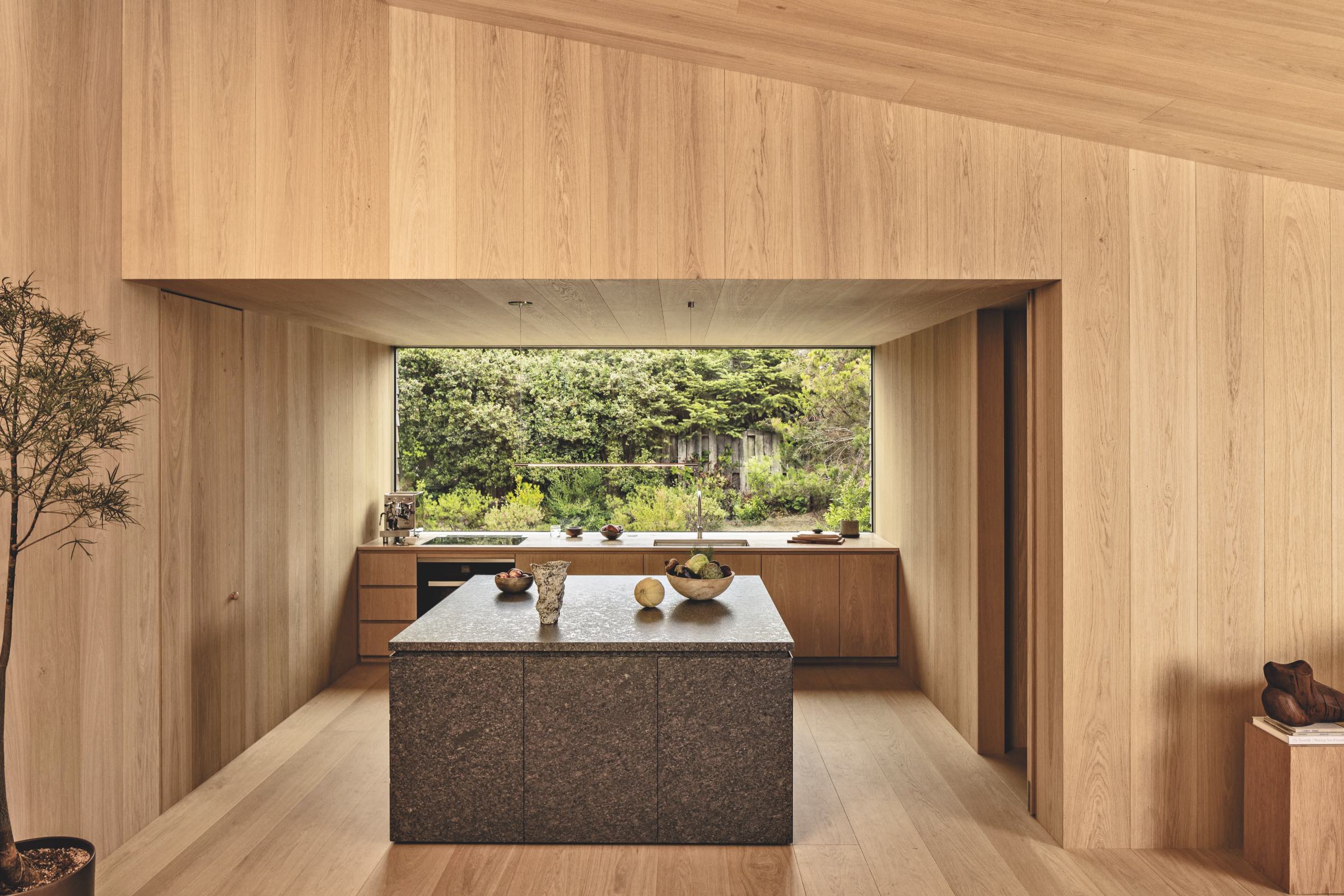 Minimalist kitchen with wood-paneled walls and ceiling, stone island countertop, and large window overlooking a green outdoor landscape.